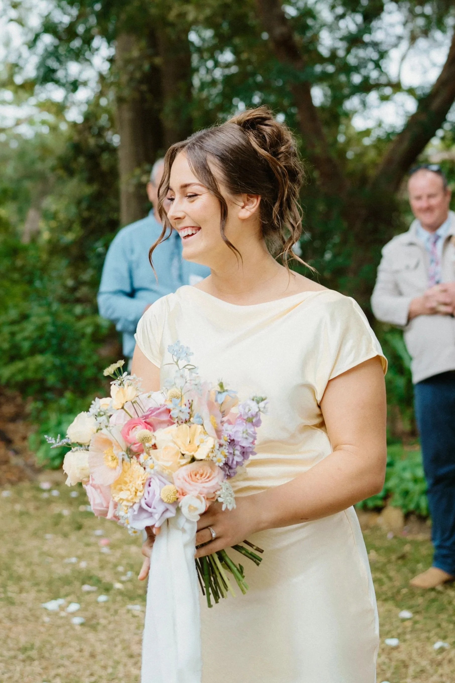 A woman in a light yellow dress holding a bouquet of pastel-colored flowers, smiling outdoors with trees in the background.