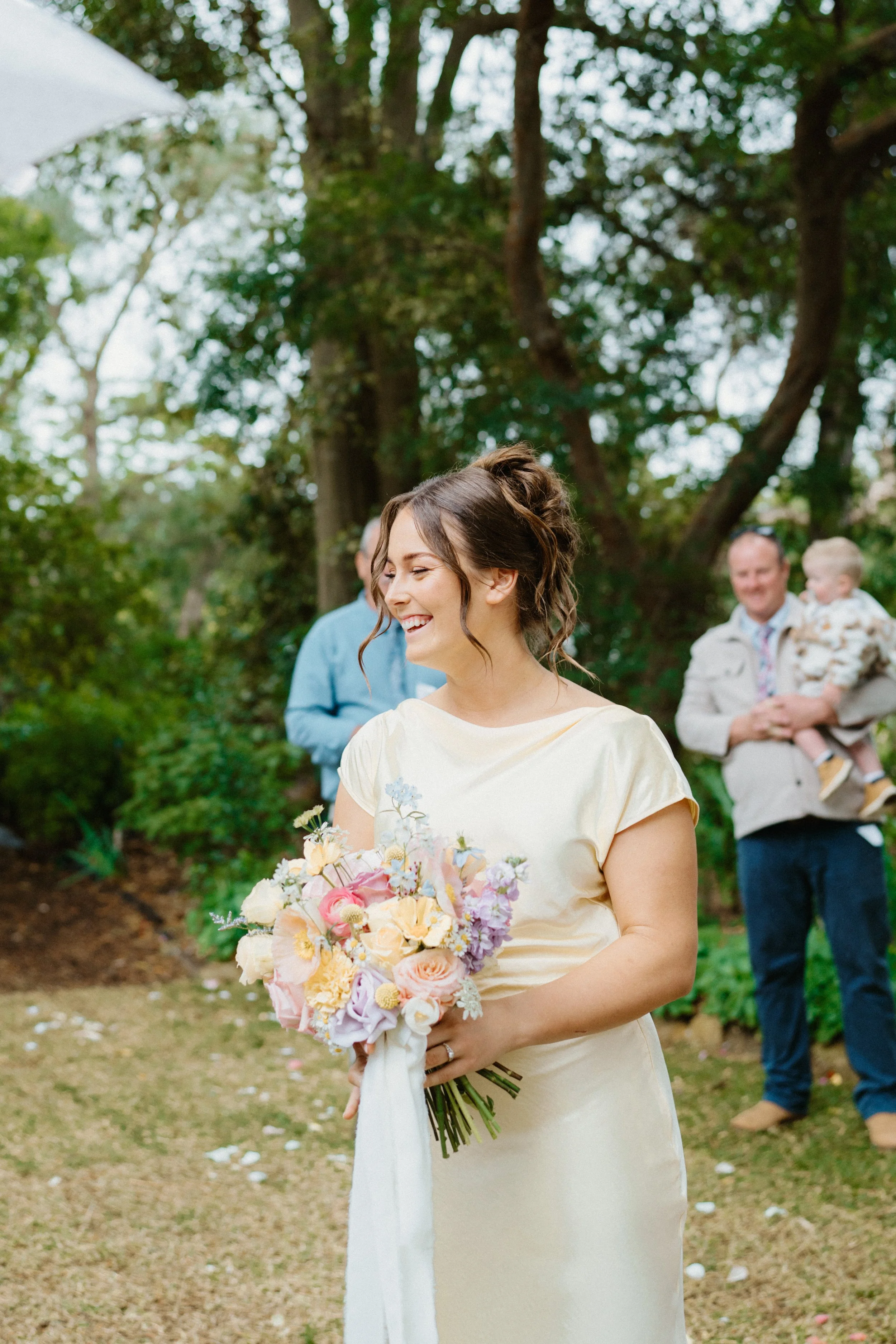 A woman in a cream-colored dress holding a bouquet of pastel-colored flowers at an outdoor wedding, smiling surrounded by trees and guests in the background.