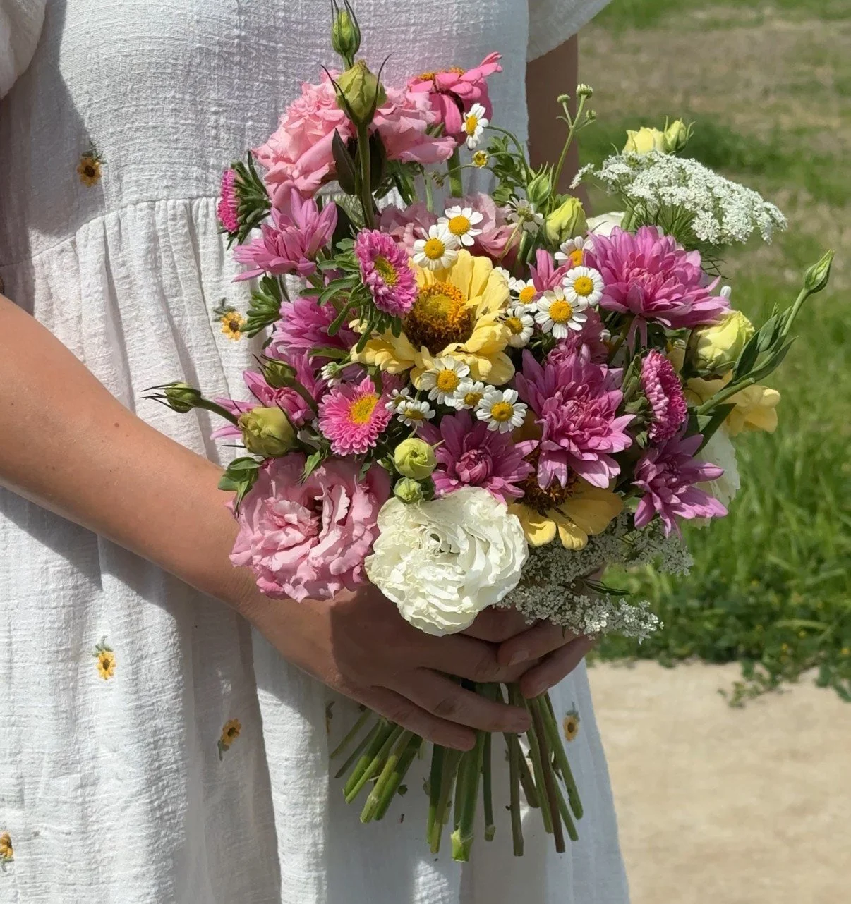 Person in a white dress holding a colorful bouquet of pink, yellow, white, and purple flowers outdoors.