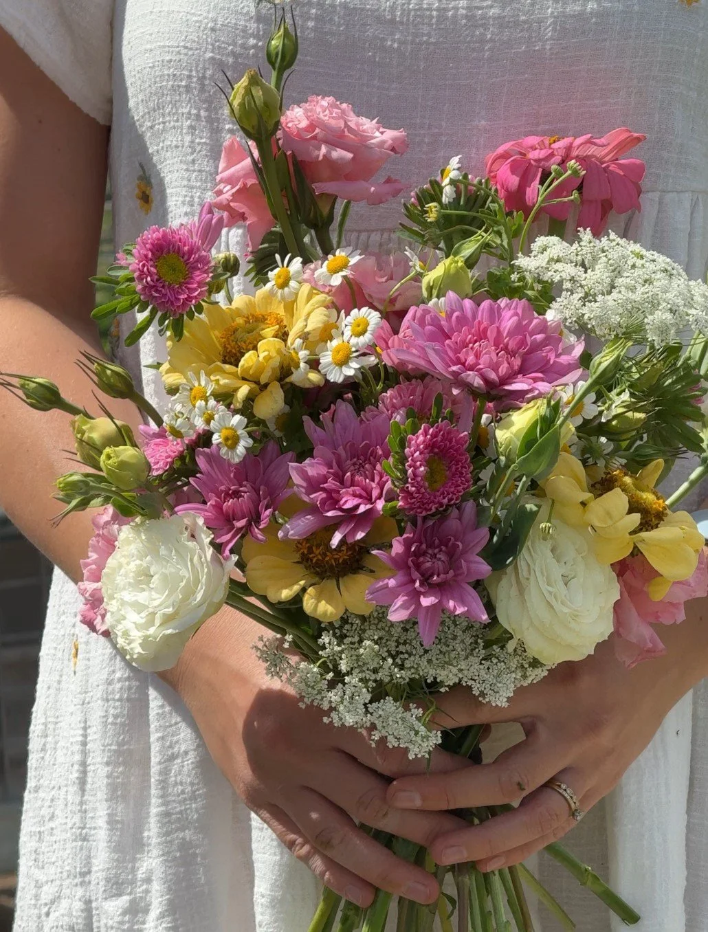 Person holding a colorful bouquet of flowers including pink, white, yellow, and purple blooms in front of a white dress.