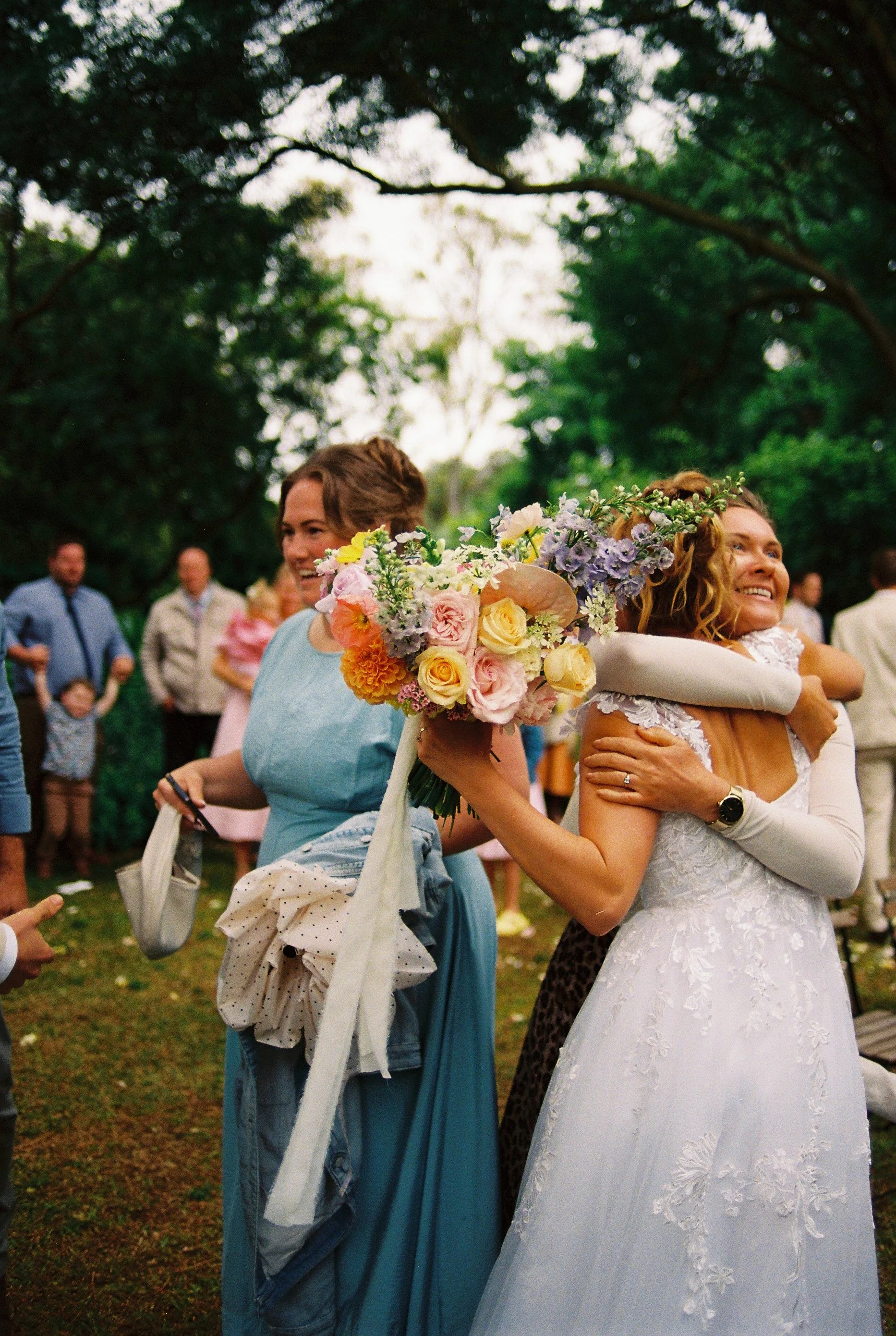 A bride with a floral crown hugging a woman holding a bouquet of flowers at an outdoor celebration.