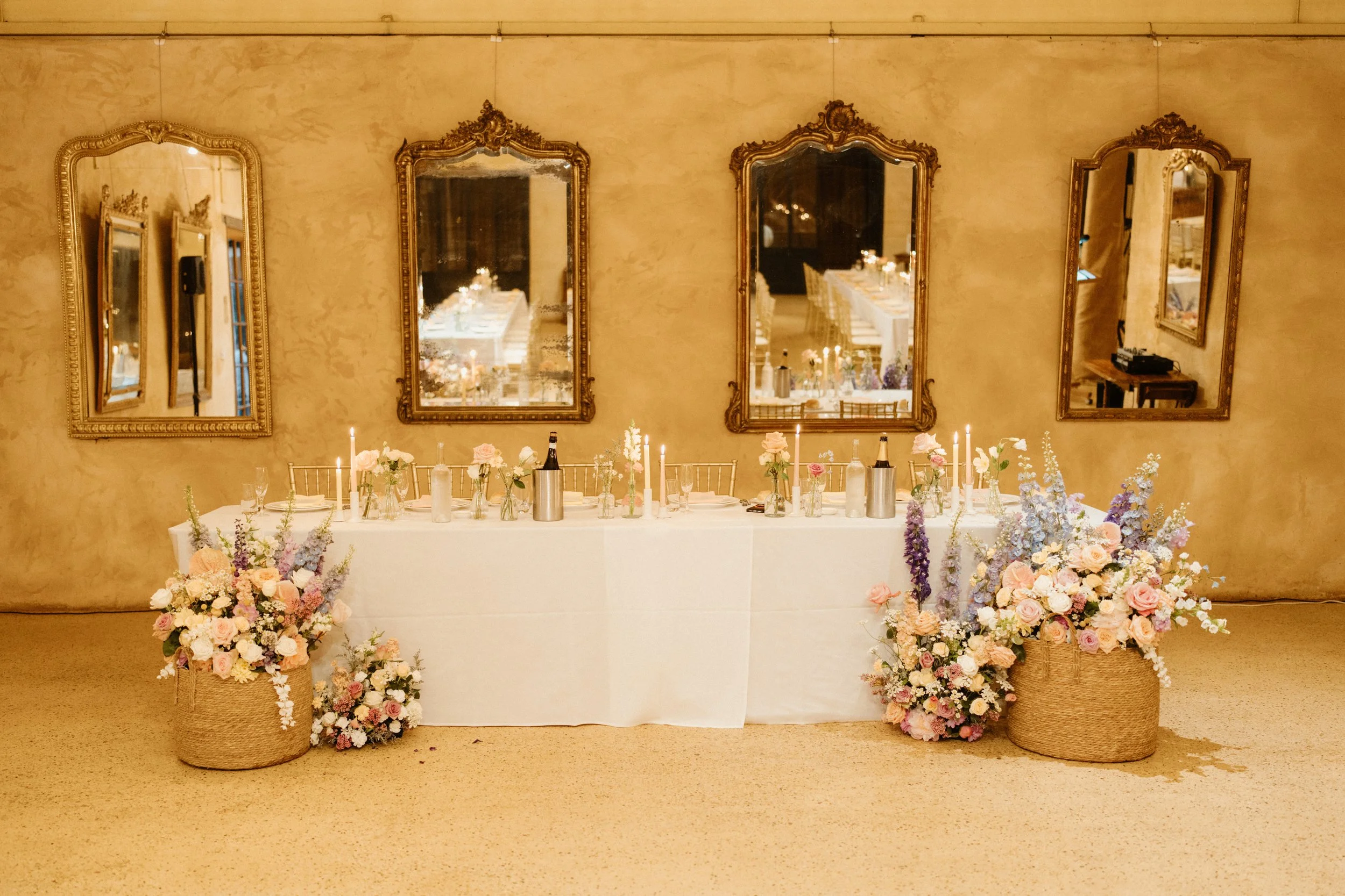 Decorated banquet table with floral arrangements, candles, and champagne bottles, set against a wall with five ornate gold mirrors.