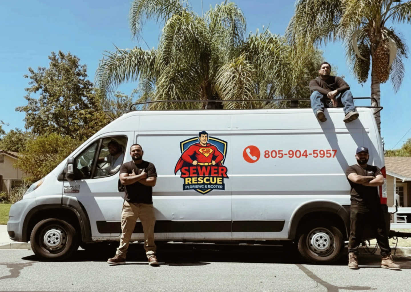 Four men posing in front of and on a plumbing and rooter service van with the company's logo and contact information, parked on a street with trees and a house in the background, under a clear blue sky.