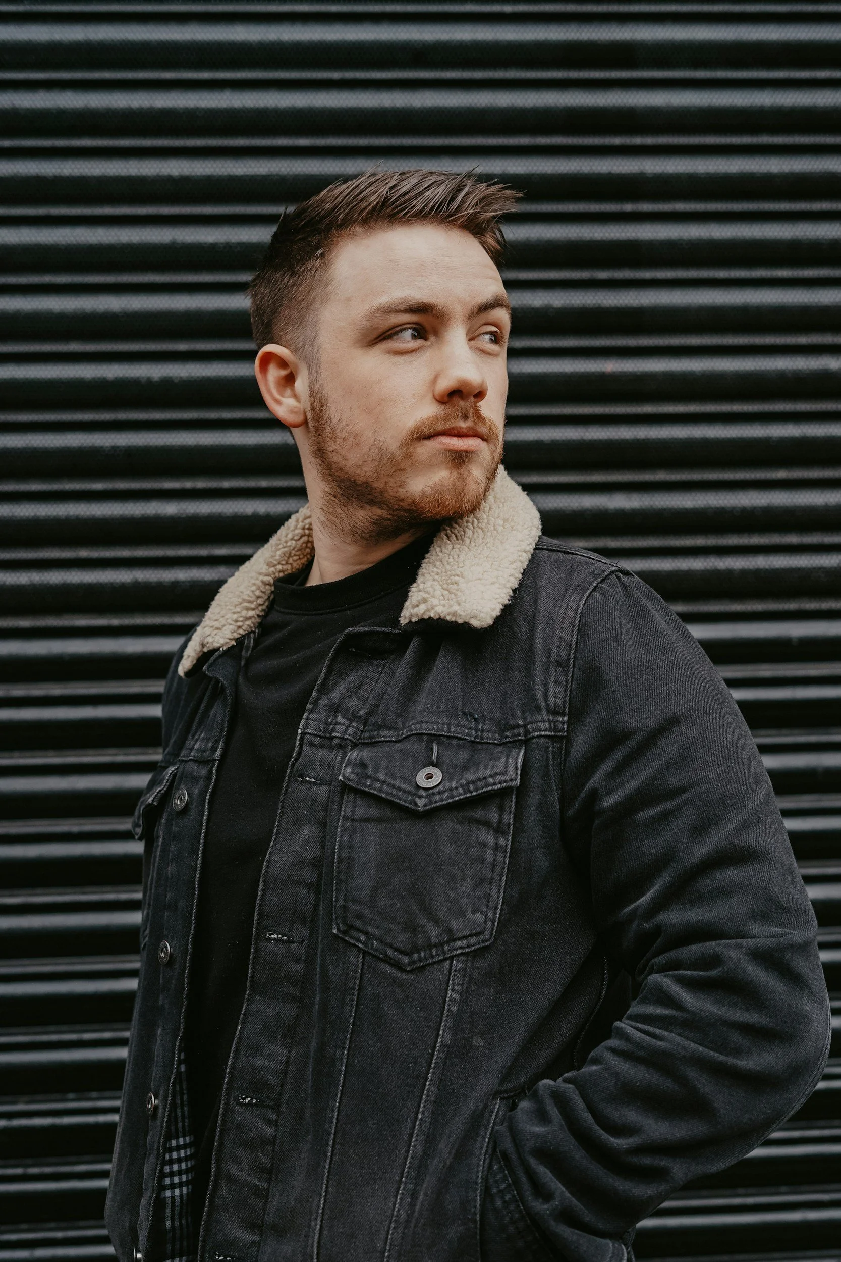 A young man with short brown hair and a beard wearing a black denim jacket with a cream-colored shearling collar standing against a black metal slat background.