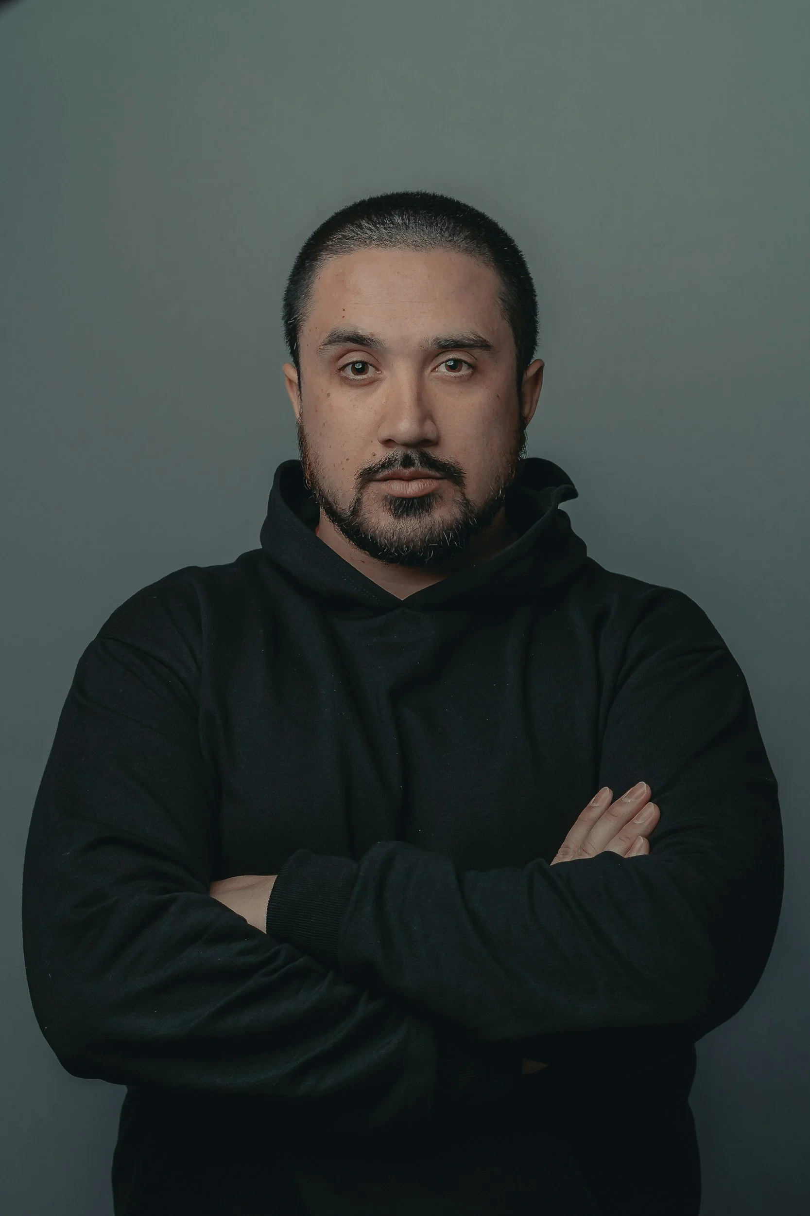A man with short black hair, a goatee, and a beard, wearing a black hoodie, standing against a plain gray background with arms crossed.
