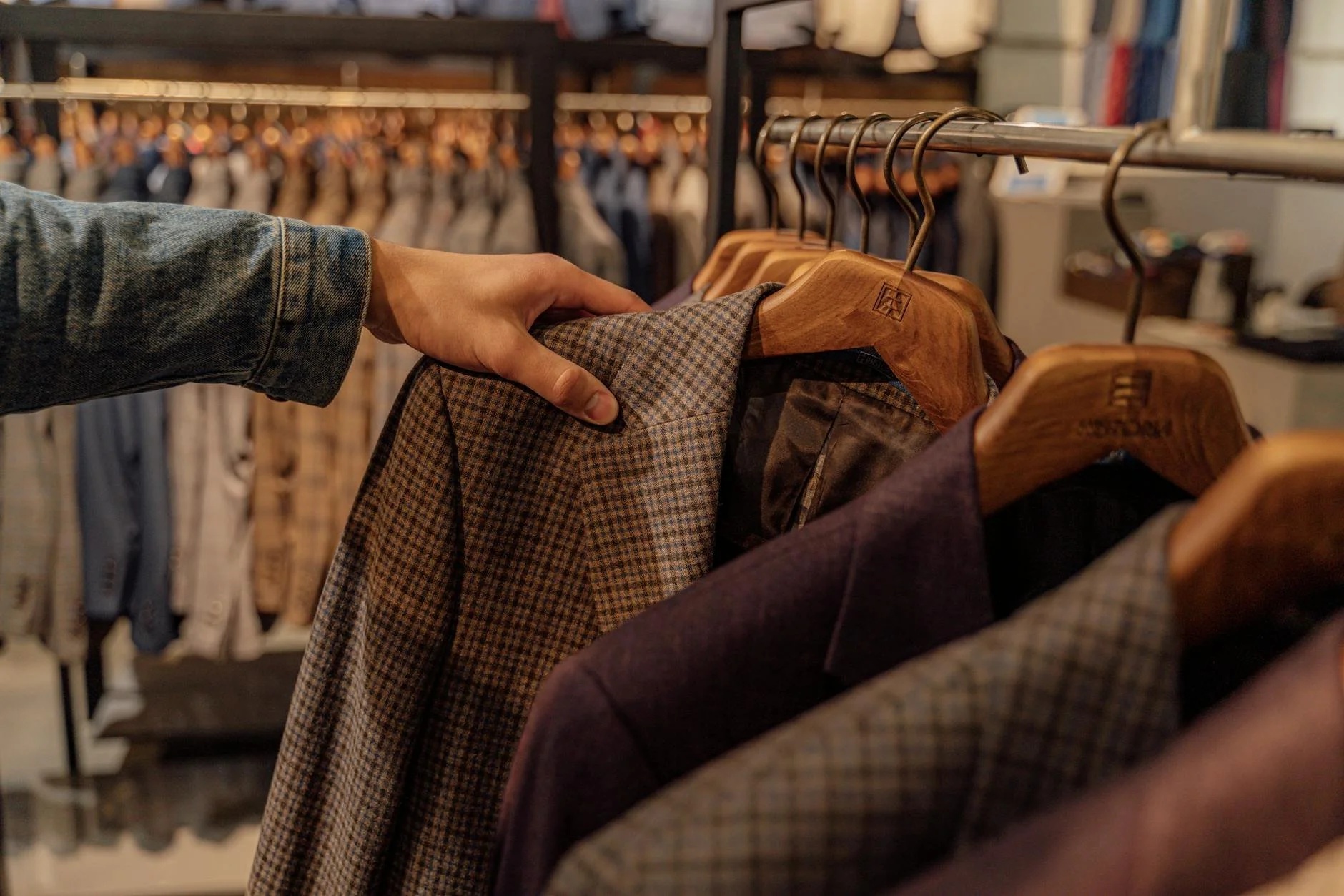 A person wearing a denim jacket shopping for men's blazers and suits at a clothing store, holding a checked blazer on a wooden hanger.