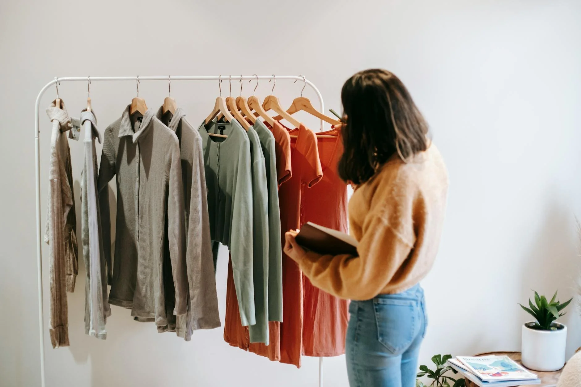 Woman browsing clothes rack with various colored tops and sweaters in a room with a plant and magazines.