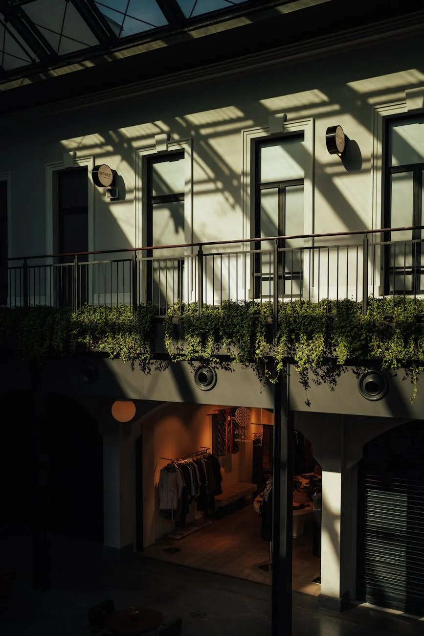 Sunlight casts shadows on the white exterior wall of a two-story building, revealing windows, a balcony with a green plant wall, and a boutique clothing store on the ground floor.