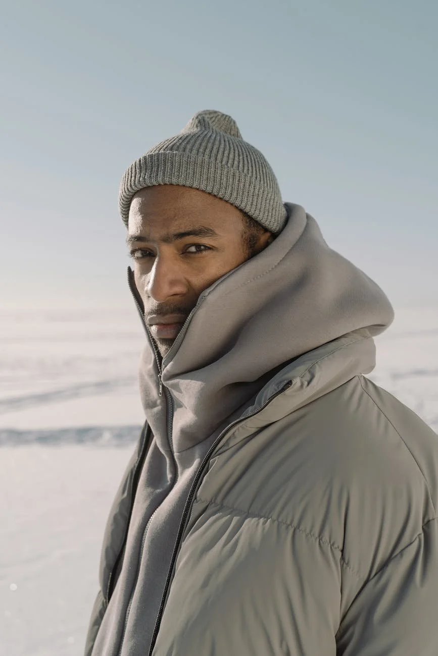 A man wearing a gray beanie and a gray puffer jacket standing outside in a snowy landscape during daytime.