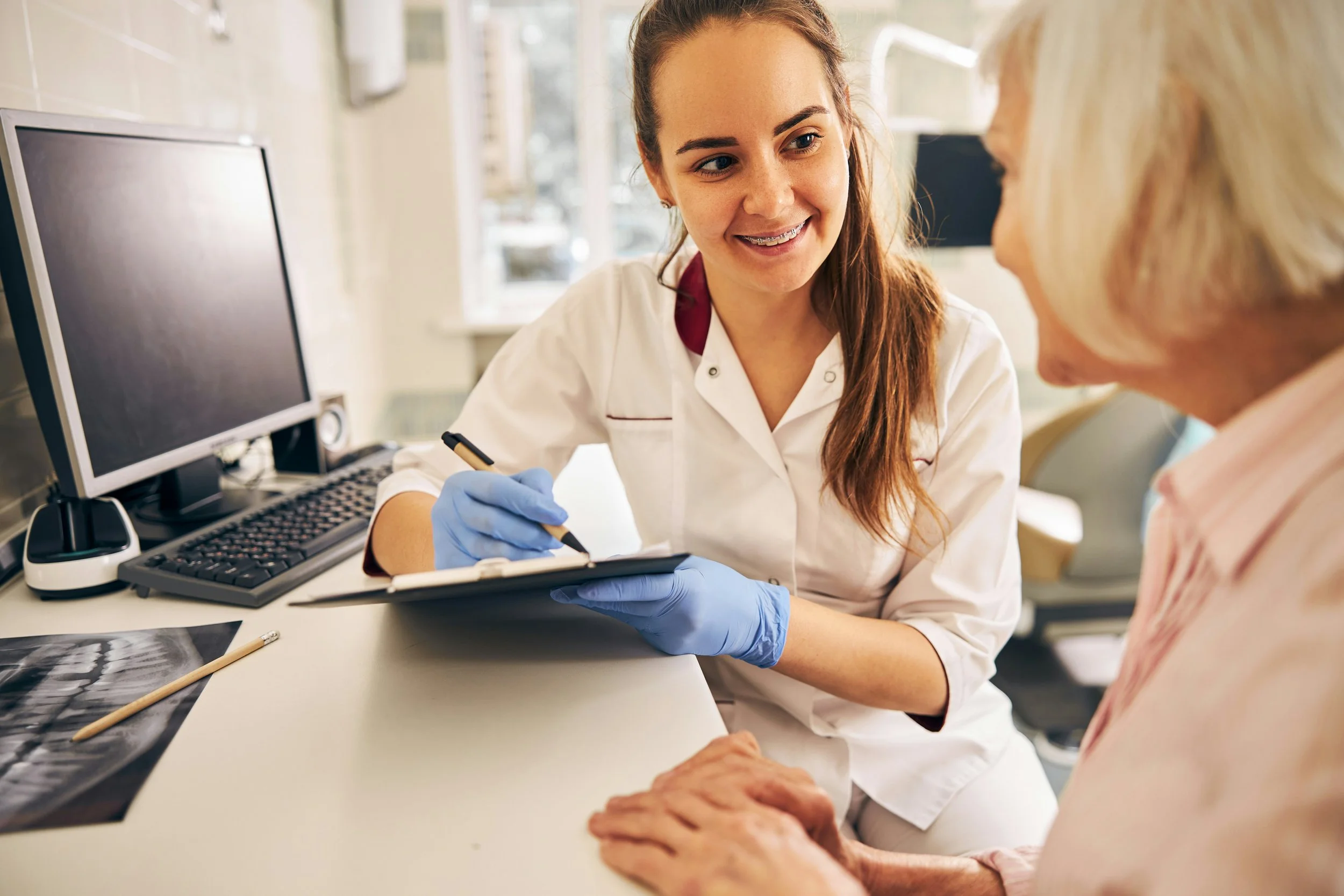 A healthcare professional, wearing a white coat and blue gloves, speaking with an elderly woman at a medical office or clinic. The professional is holding a clipboard and smiling, with medical images, a computer, and office supplies on the desk.