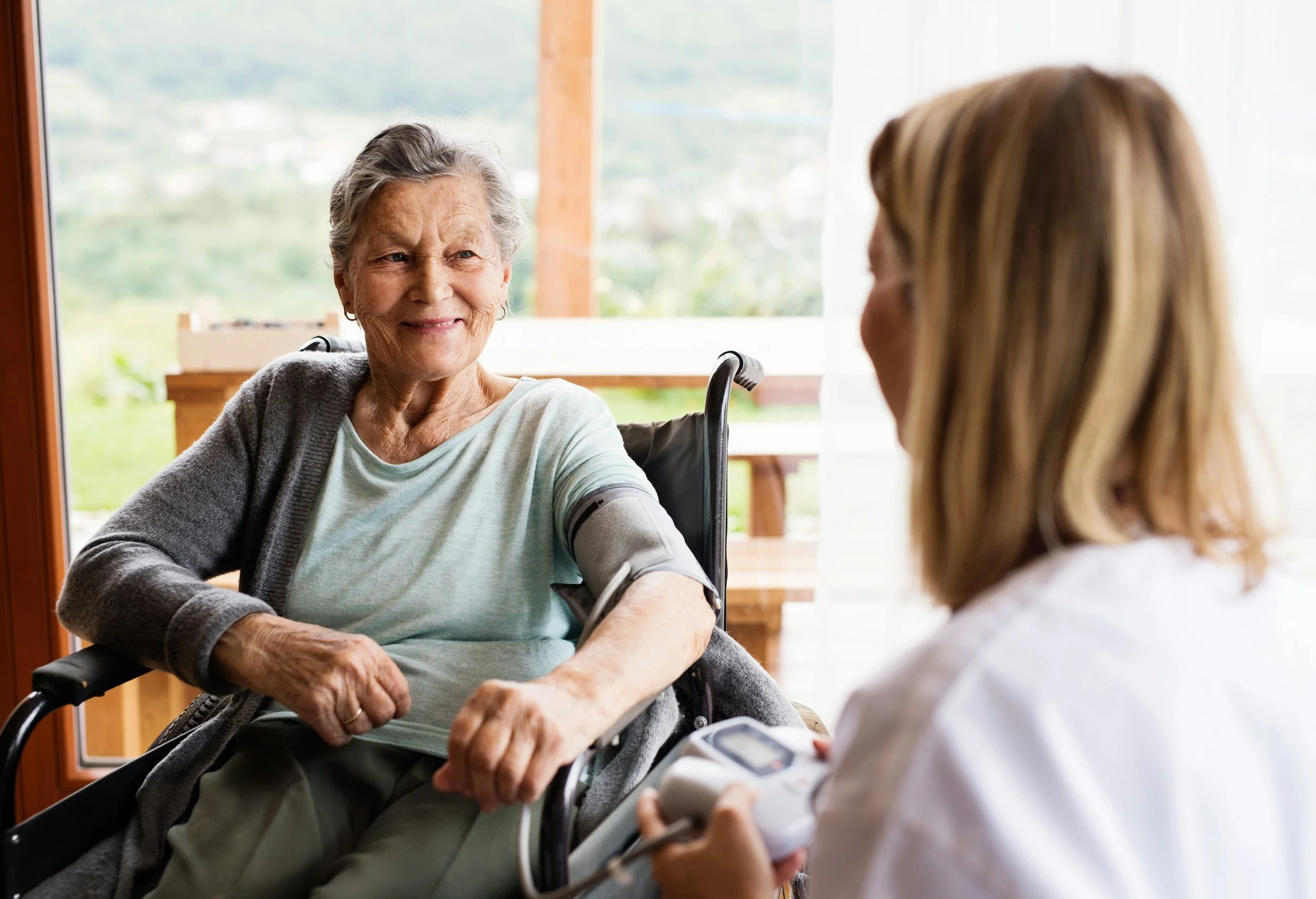 A senior woman sitting in a wheelchair, smiling at a nurse taking her blood pressure in a bright room with a view of the outdoors.