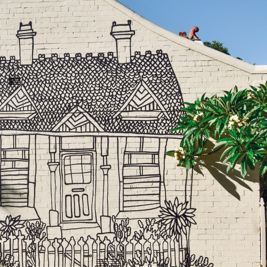 A mural of a house drawn in black lines on a white brick wall, with a real green plant and flowers in front.
