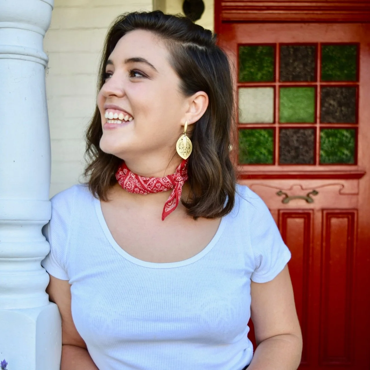 A young woman with shoulder-length dark hair smiling, wearing gold earrings, a white t-shirt, a red bandana around her neck, standing near a white pillar with a red decorative door in the background.