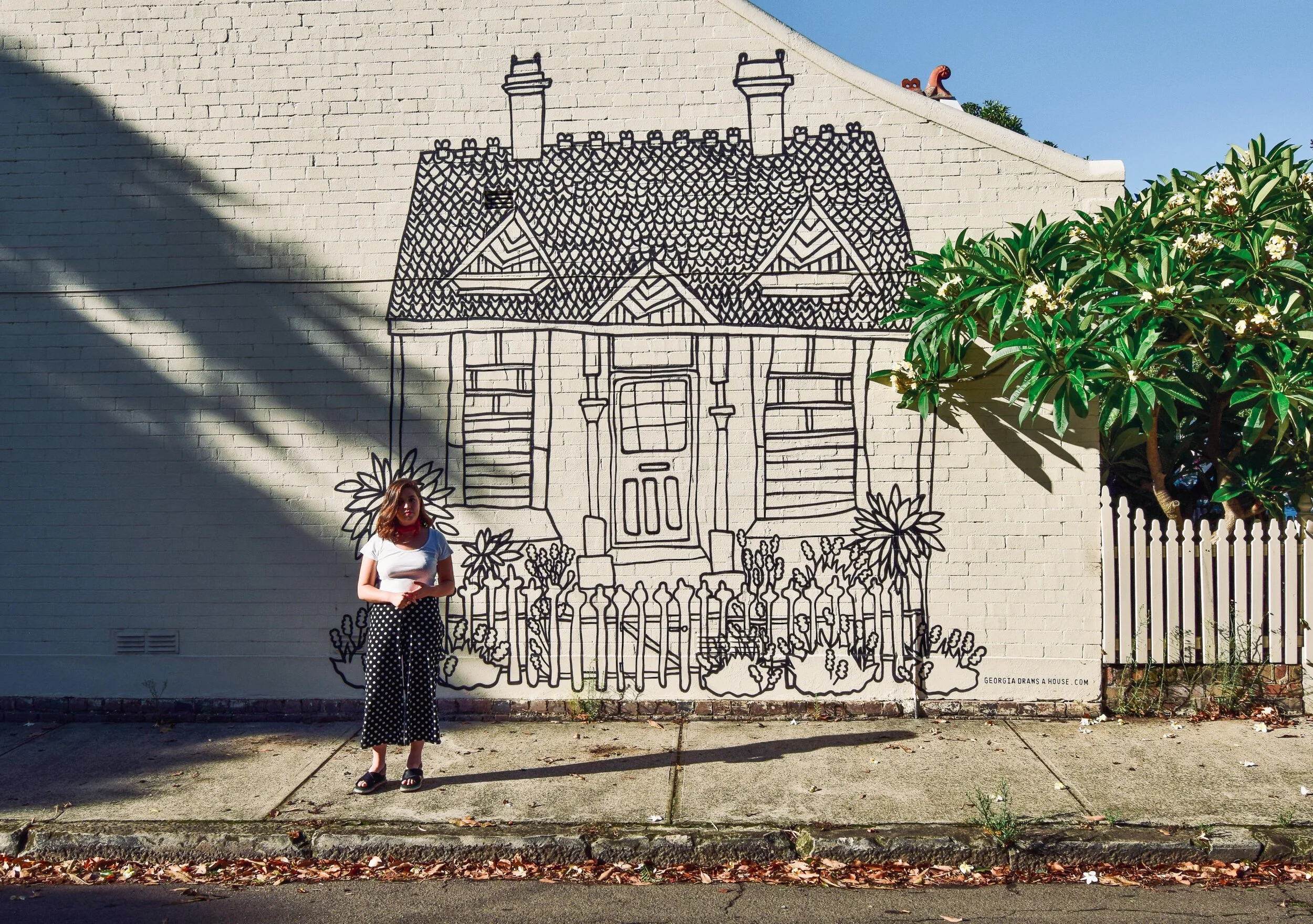 A woman standing in front of a mural of a house drawn on a brick wall. The mural features a simple house with a fence and plants in front. The woman is wearing polka dot pants, a white top, and has a red scarf around her neck.