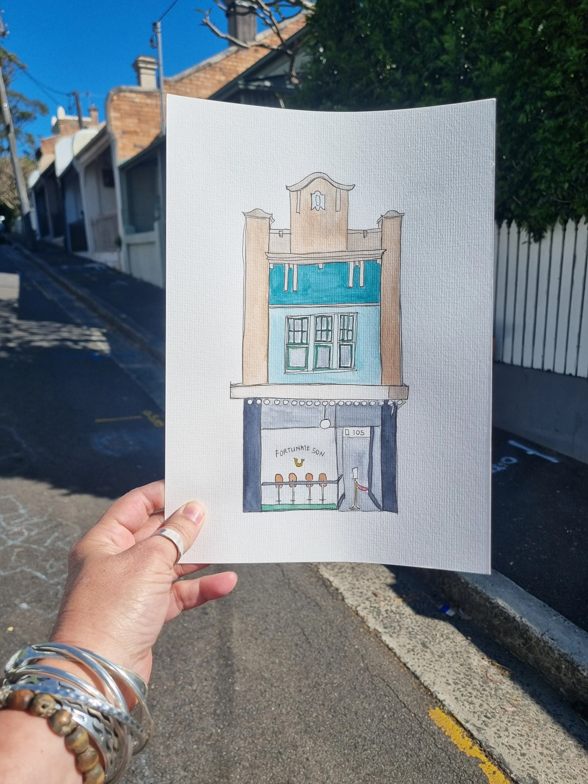 A hand holding a drawing of a three-story building with a blue and beige facade, a balcony with blue curtains, and a storefront with a sign that says "Fortunate Son." The building is on a street with houses and trees in the background.