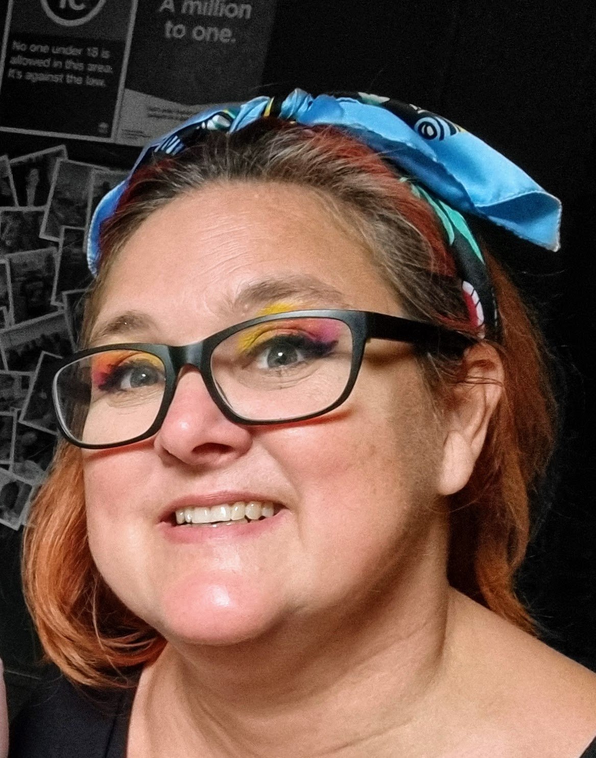 A close-up of a woman with short, reddish hair, wearing black glasses with colorful eye makeup, including pink, yellow, and orange eyeshadow. She has a blue patterned headscarf and a black top, and is smiling slightly. The background contains black-and-white photo displays and a poster that reads 'A million to one.'