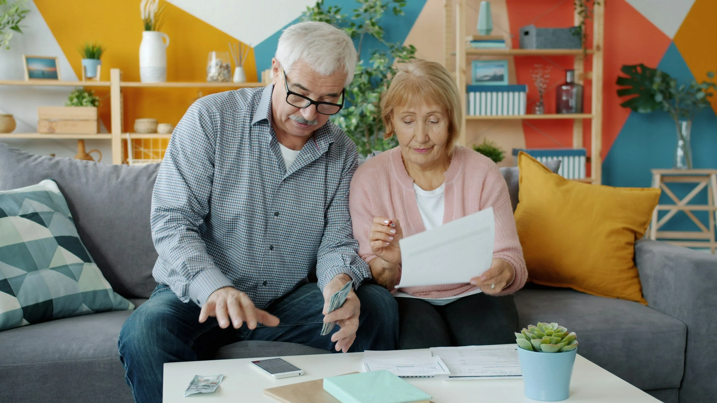 An elderly couple sitting on a gray couch in a colorful living room looking at documents.