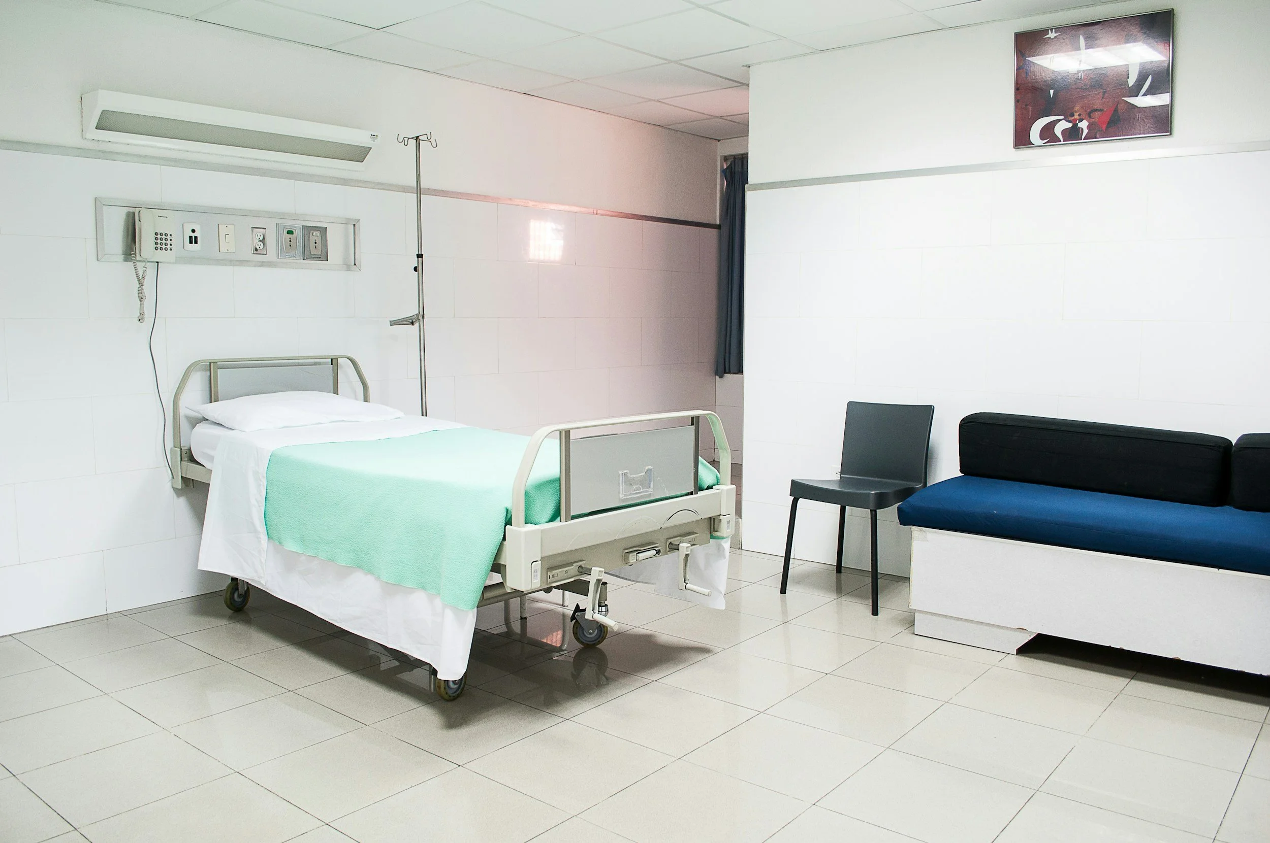 Empty hospital room with a hospital bed, a chair, a sofa, and medical equipment.