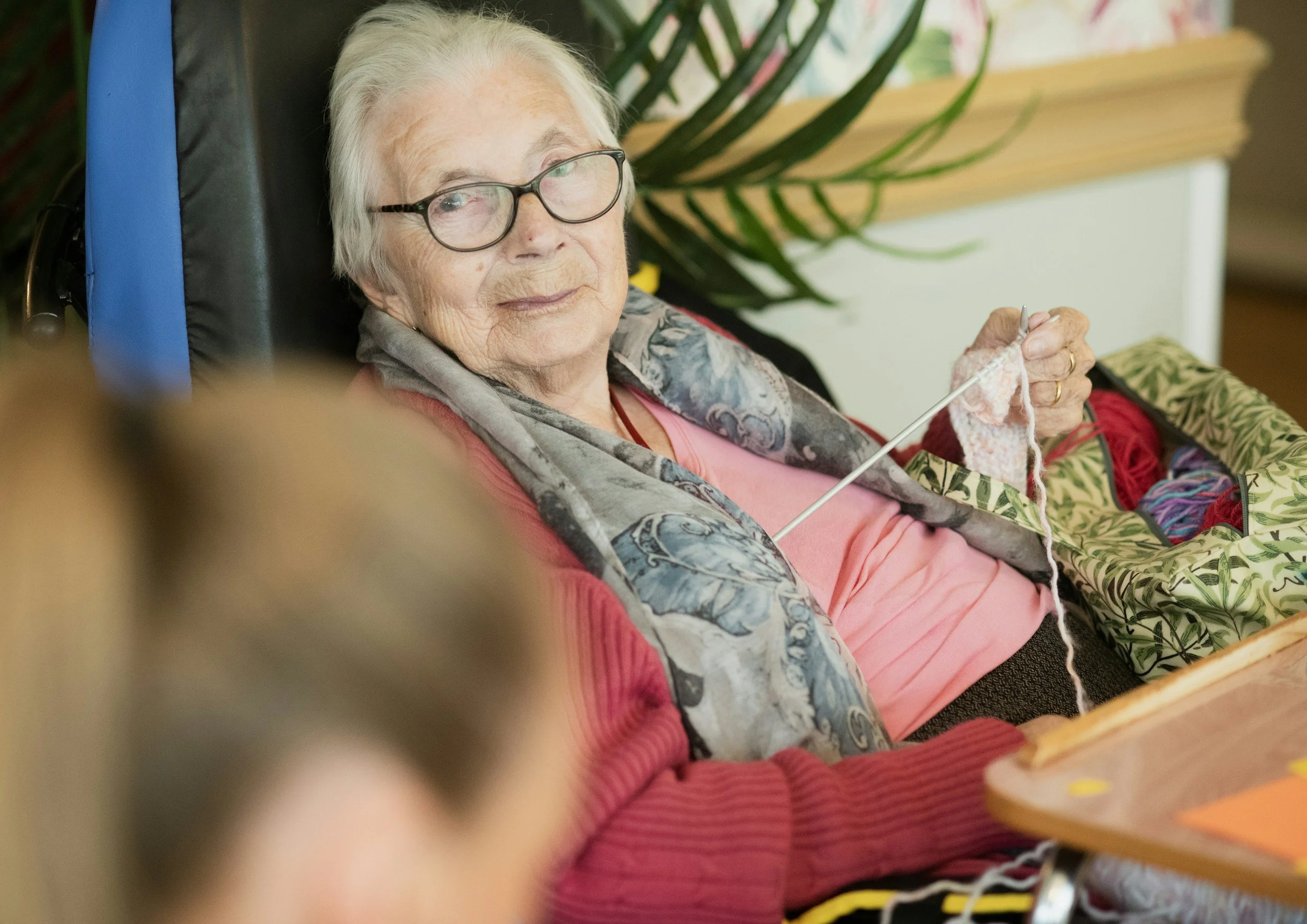 An elderly woman with white hair and glasses sitting in a wheelchair, knitting with pink yarn, with a patterned blanket nearby, in a room with plants and colorful decor.