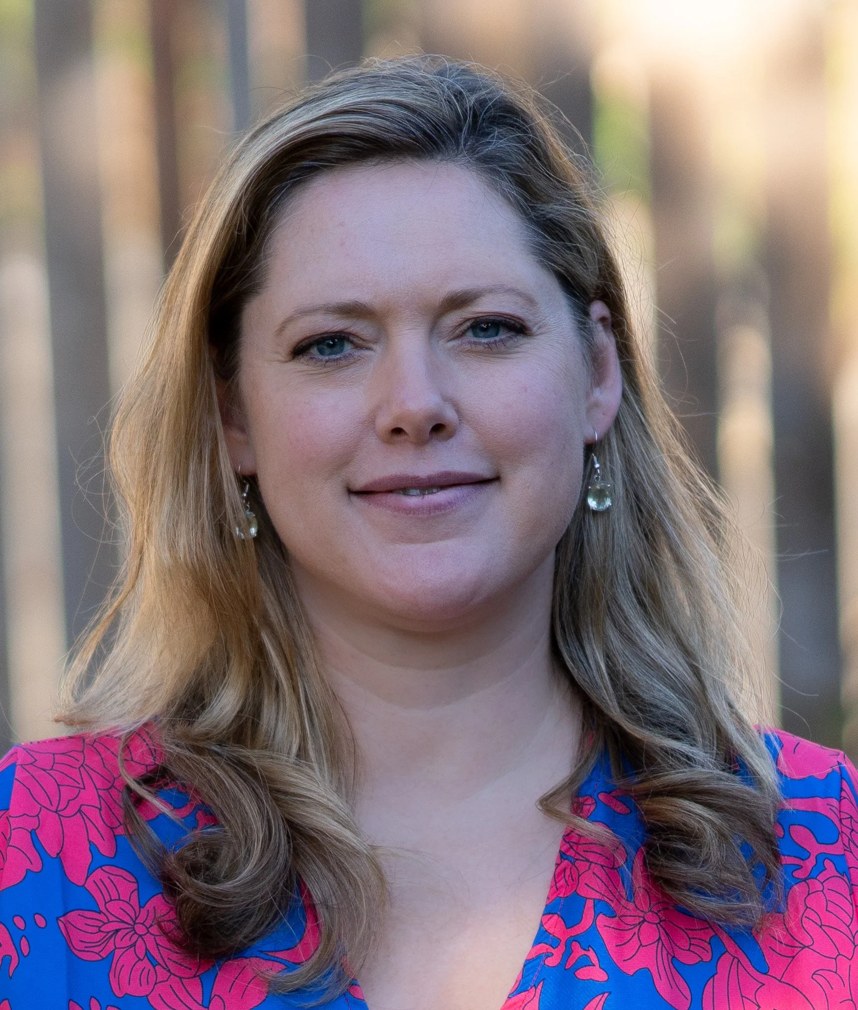Close-up of a woman with blue eyes and wavy blonde hair, wearing a pink and blue patterned shirt and earrings, outdoors with blurred background.