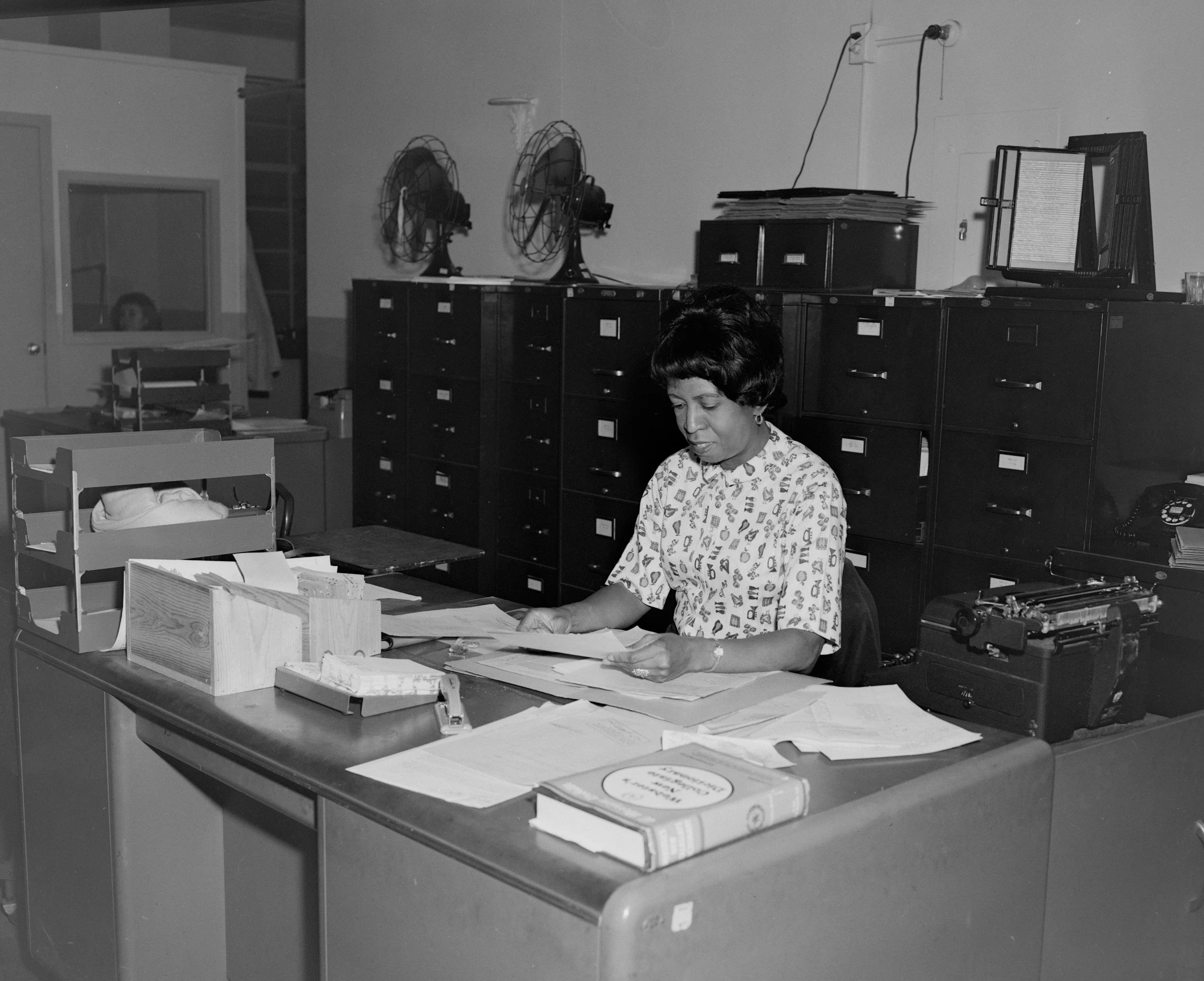 Vintage black and white photo of a woman working intently at a busy office desk.