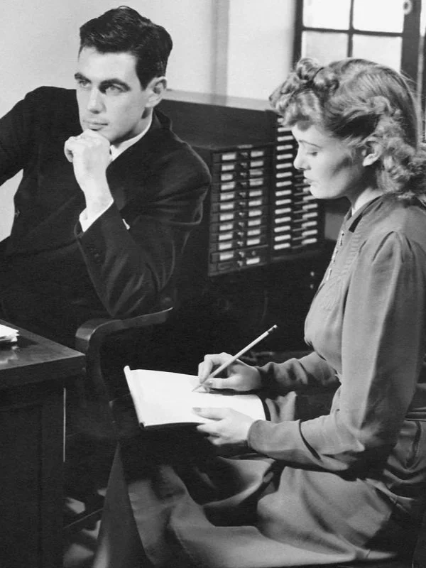 Black and white vintage photo of a man and woman in the office, he's dictating notes to her.