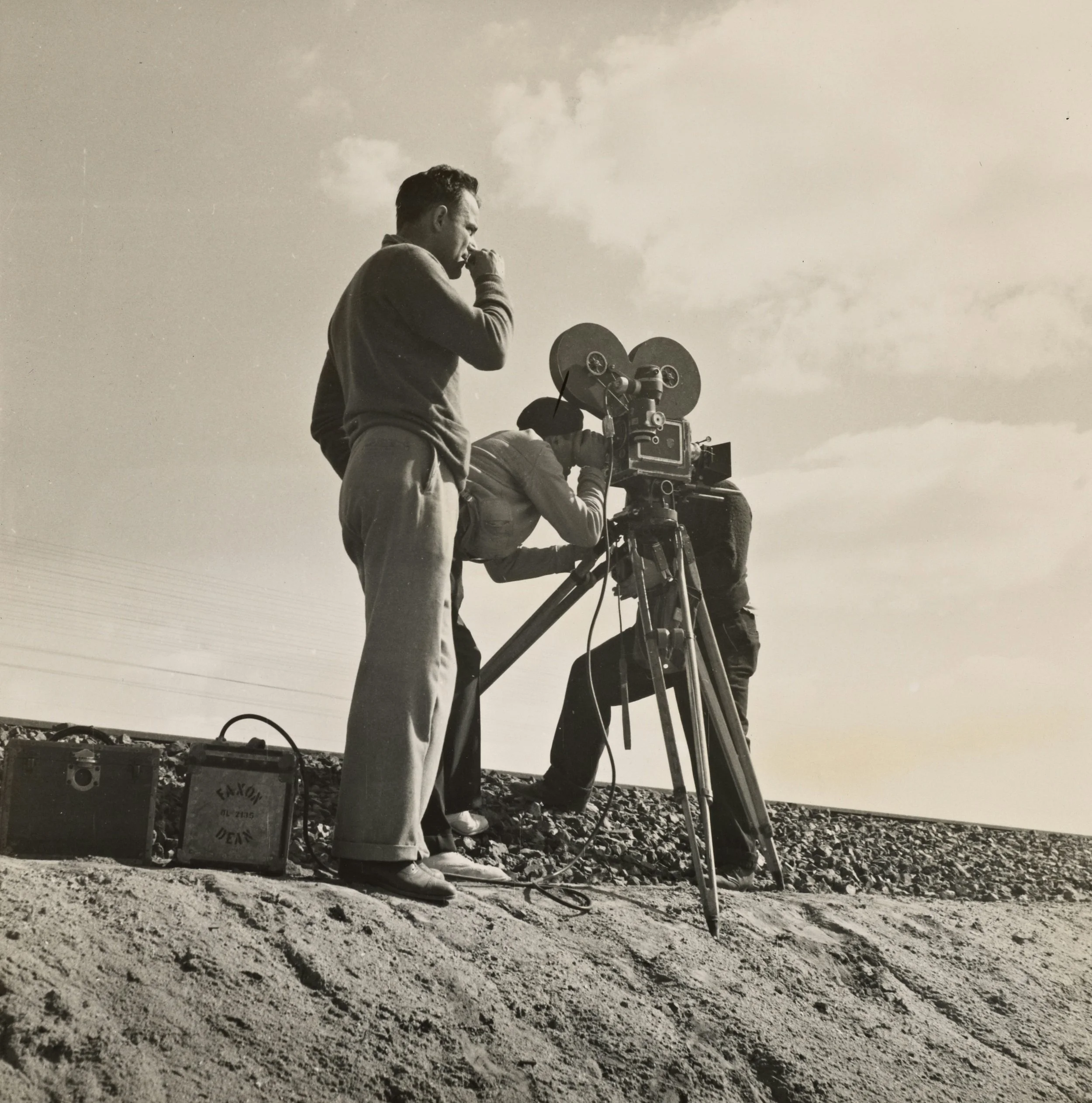 Black-and-white photo of a man operating a vintage film camera on a tripod with others nearby.