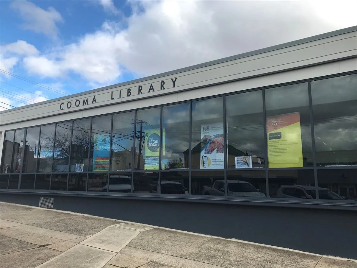 Exterior of the Cooma Library with large glass windows reflecting the cloudy sky and parked cars outside.