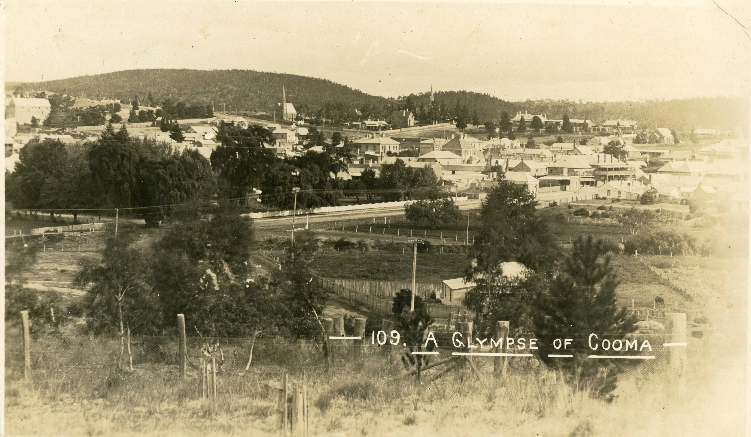 An old sepia-toned photograph of Cooma, NSW with houses, church steeples, and a school or large building in the foreground, with hills in the background. The caption reads '109. A Glympse of Cooma.'