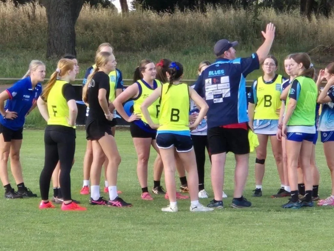 A group of young female athletes listening to their coach during a sports practice on a grassy field.
