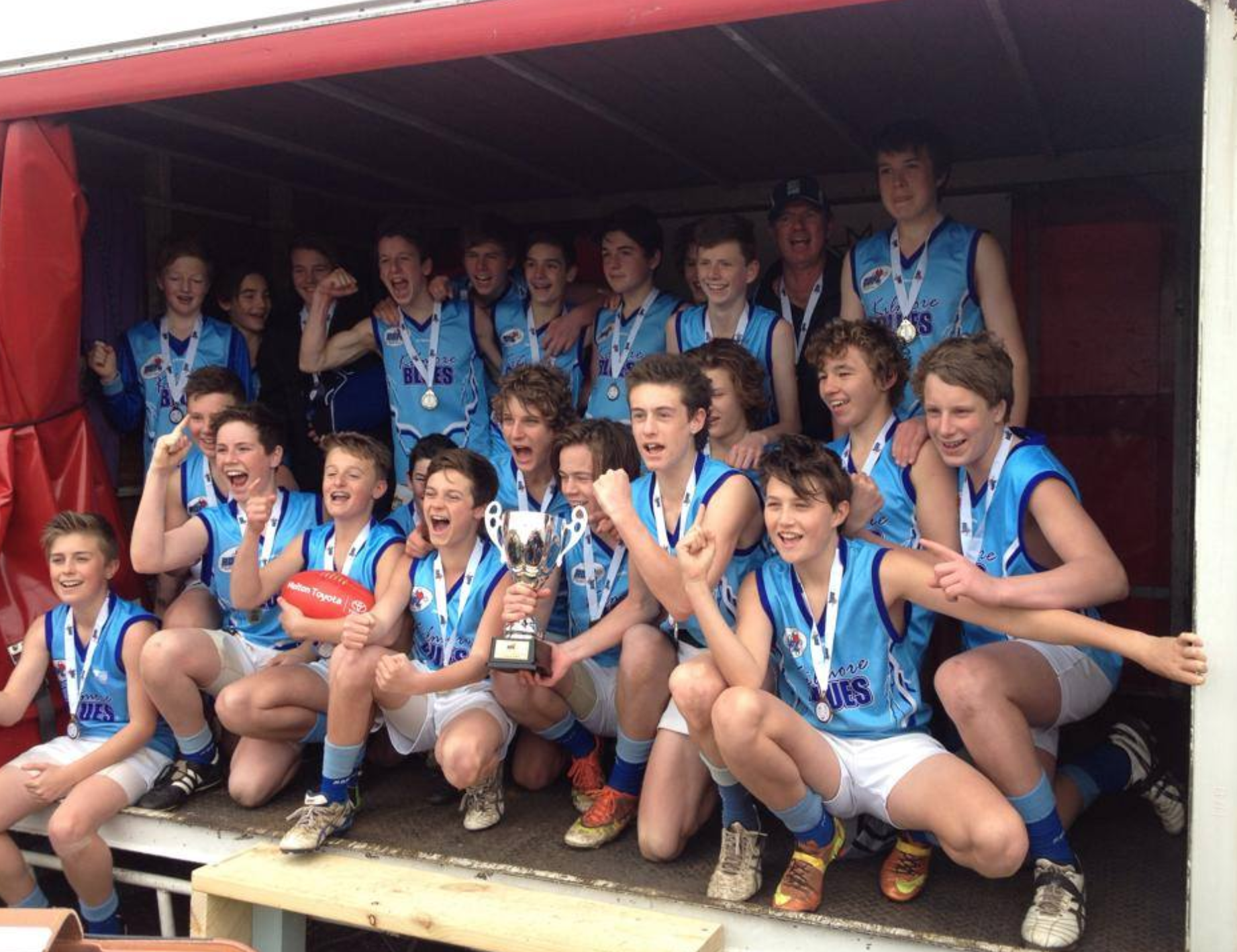 A group of junior football players and a coach celebrating a victory on a sports team, with some holding medals and a trophy, all wearing blue sports jerseys and shorts.