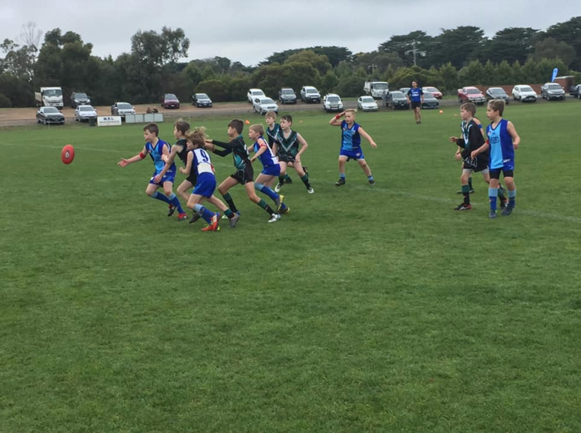 Kilmore junior football team playing on a grassy field with cars parked in the background.