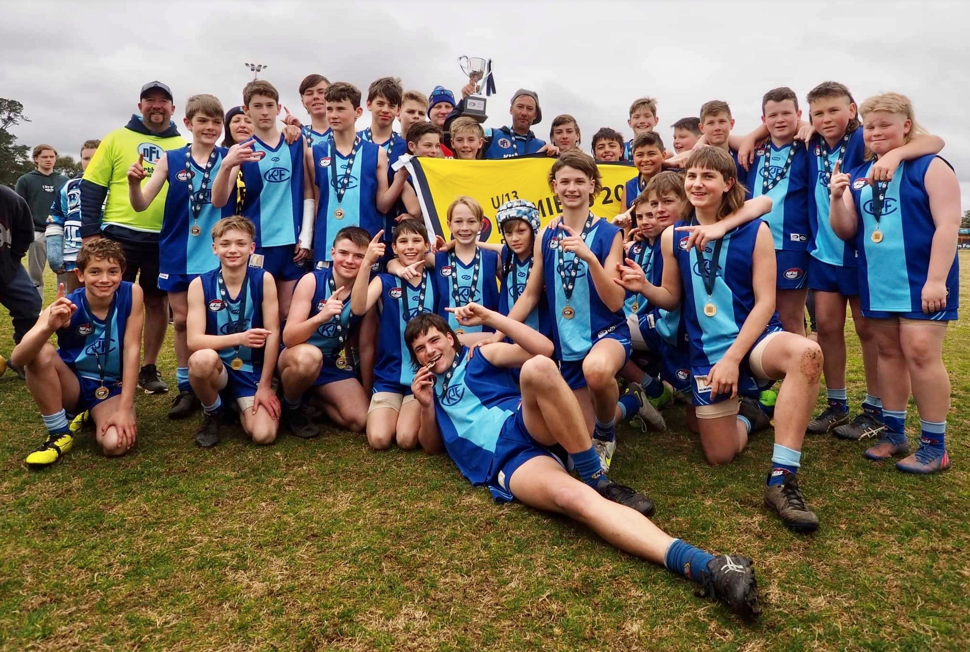 Kilmore junior football team celebrating victory on the field, some kneeling and others standing, wearing blue uniforms with medals, holding a trophy and a yellow banner.