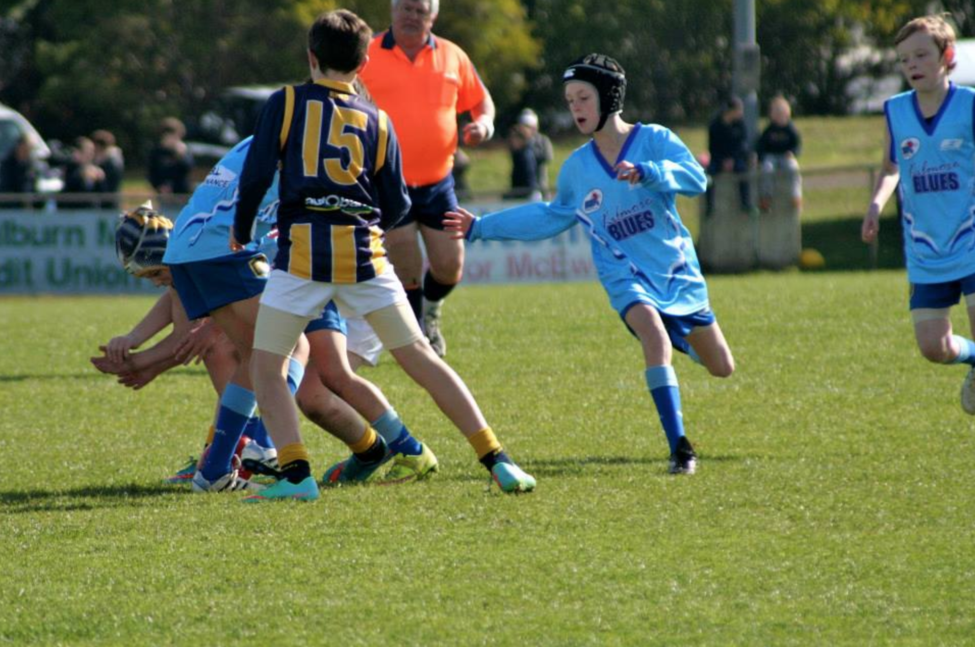 Kilmore junior football team playing on a green field, wearing blue and yellow uniforms, with a referee supervising in the background.
