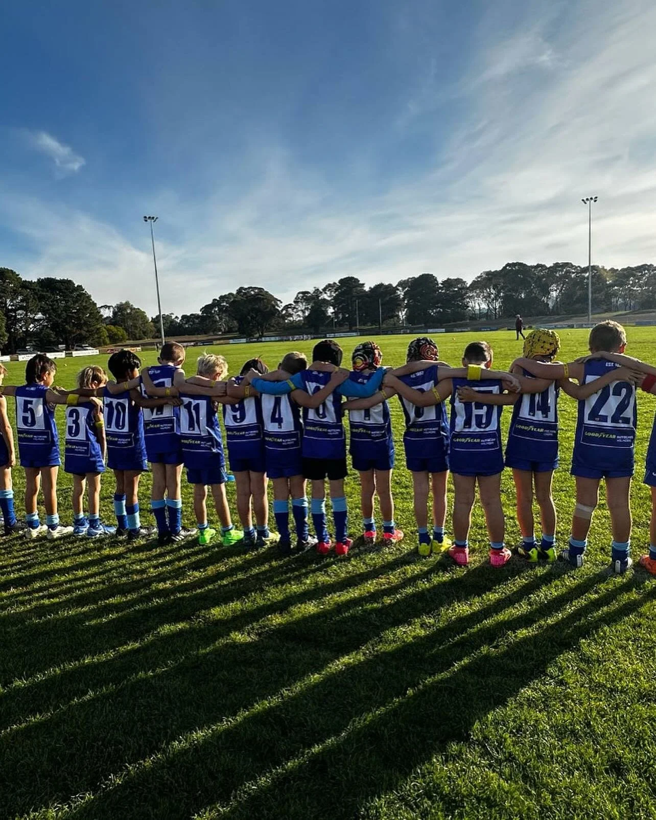 Kids in blue sports uniforms standing in a line on a soccer field with arms around each other, preparing for a game under a clear blue sky.