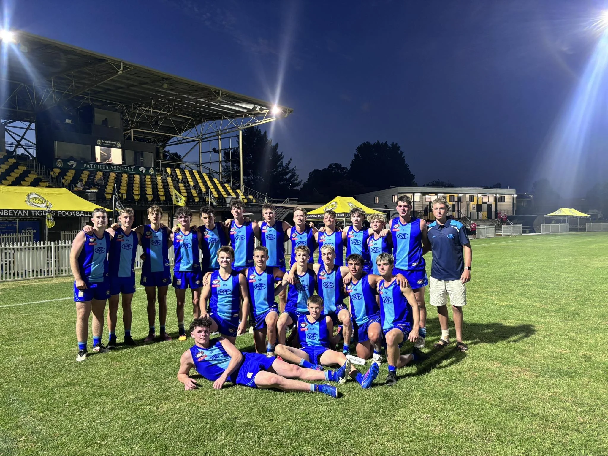 A group of young male athletes wearing blue sports uniforms, some kneeling and some standing, posing for a team photo on a sports field at night, with a stadium structure and trees in the background.