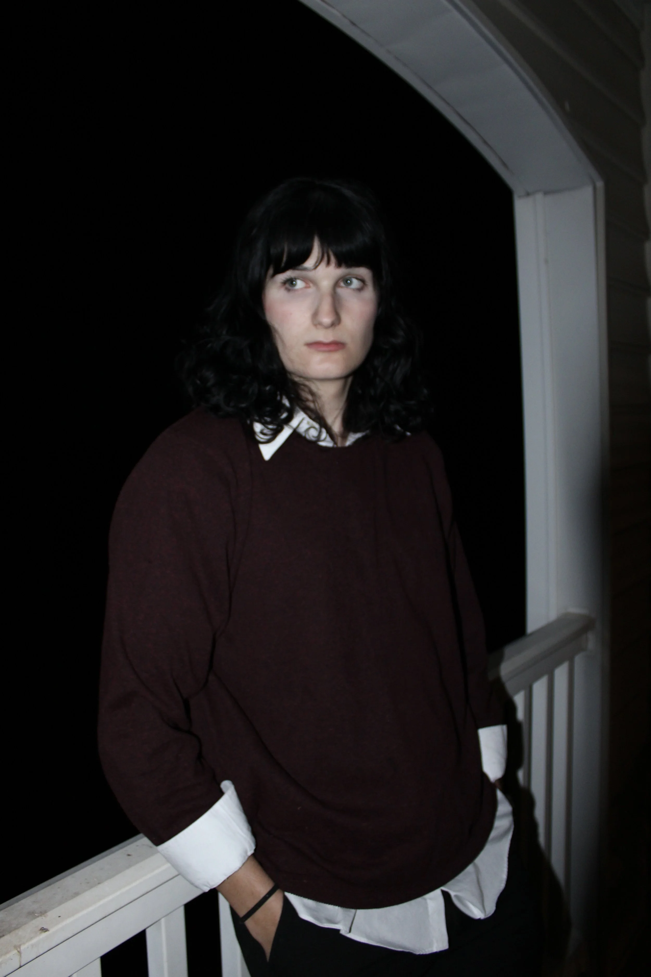 A woman with black curly hair and fair skin stands on a porch at night, wearing a dark maroon sweater over a white collared shirt, with her hands in her pockets, looking to the side.