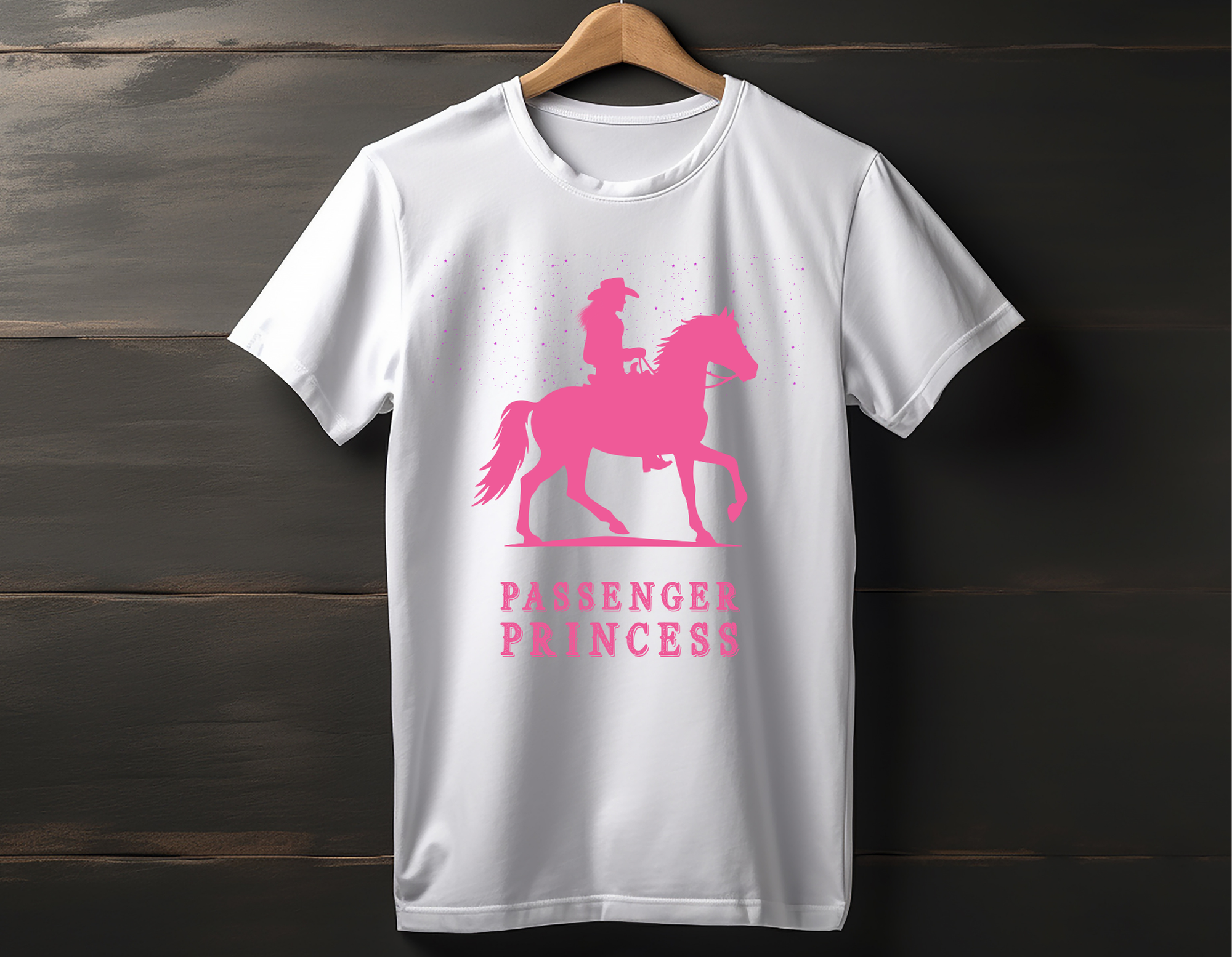 White T-shirt with pink graphic of a girl riding a horse and the words "Passenger Princess" underneath, hanging on a wooden background.