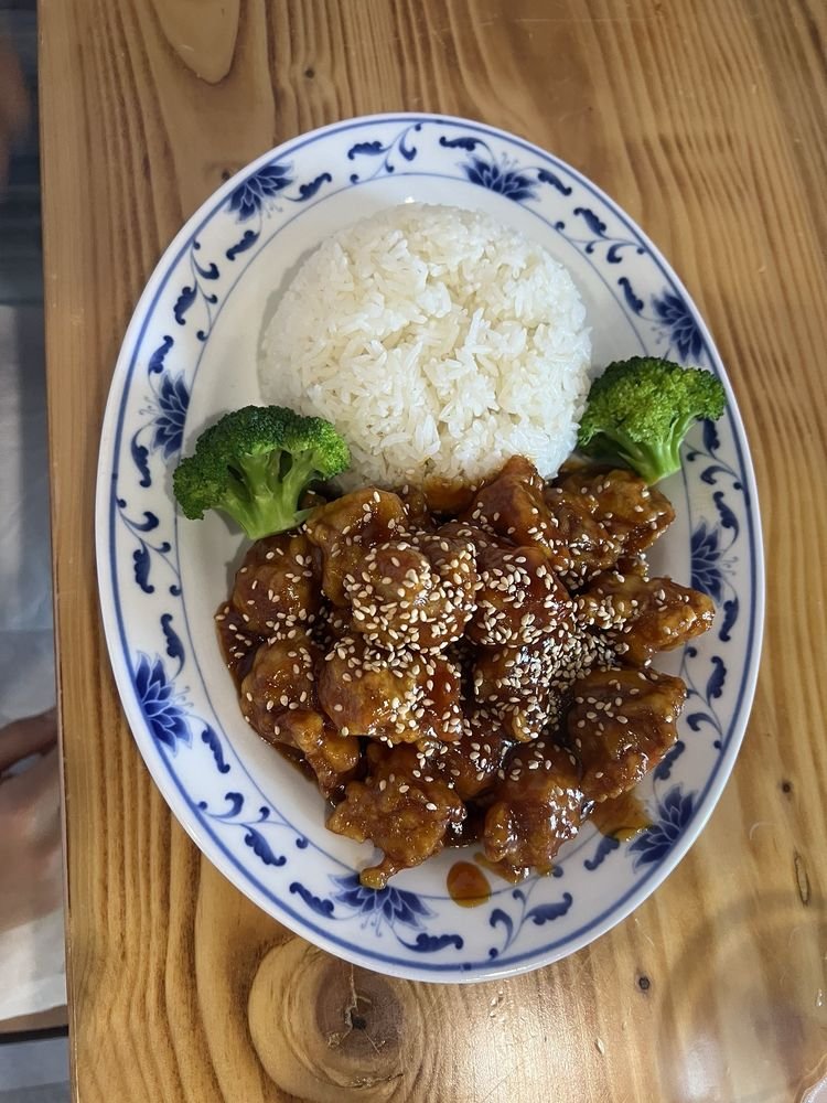Plate of Chinese food with white rice, sesame chicken, and broccoli on a wooden table.
