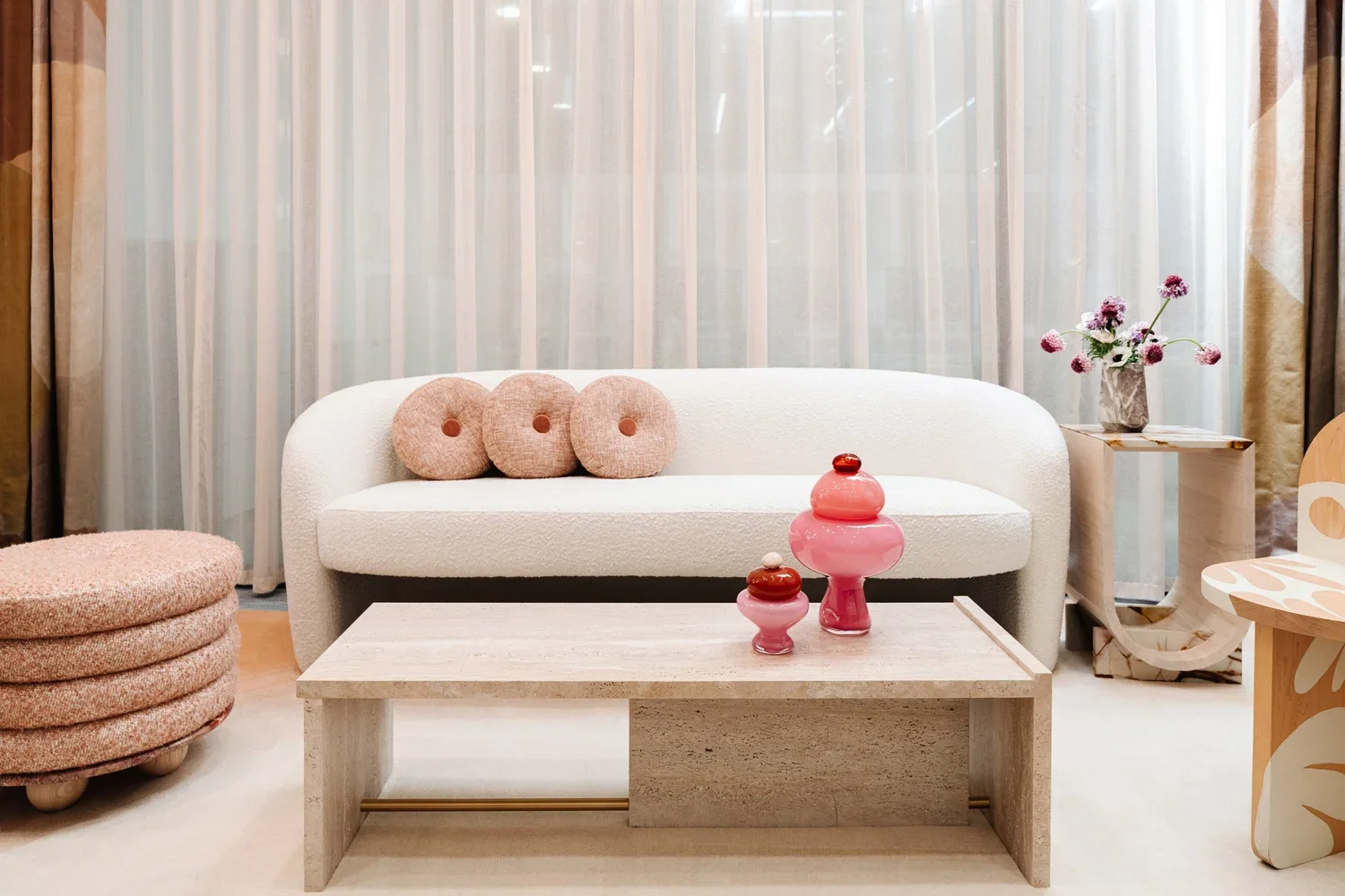 Living room with a white sofa, three round pink cushions, a beige table with pink and red vases, a side table with a vase of pink flowers, curtain background, and a patterned chair.