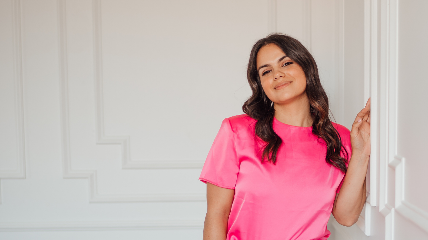 A woman with dark hair wearing a bright pink satin blouse, standing by a white wall, smiling softly.