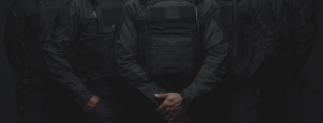 A group of people dressed in black tactical clothing standing close together, with one person clasping their hands in front of them.