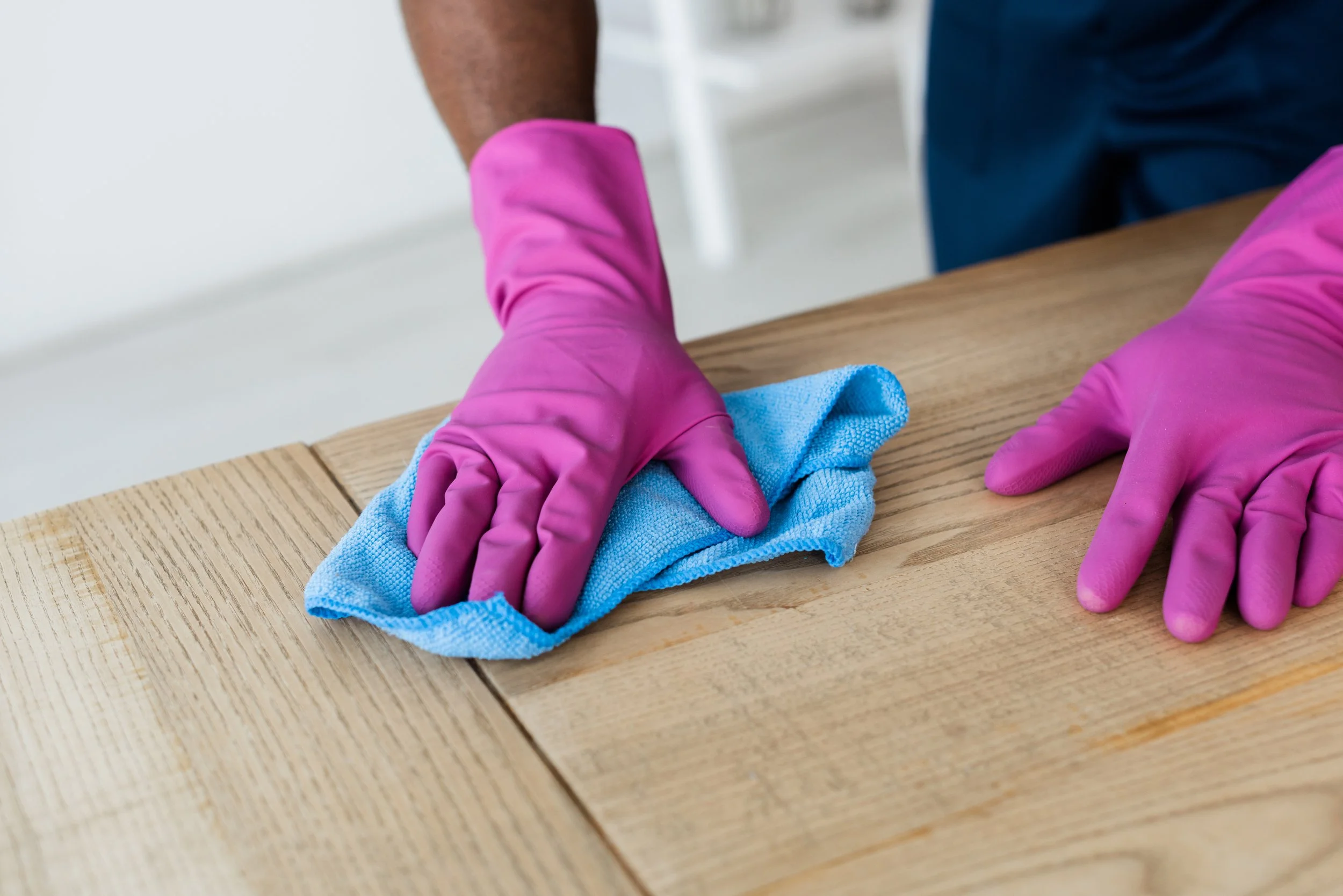 Person wearing pink gloves cleaning a wooden table with a blue cloth.