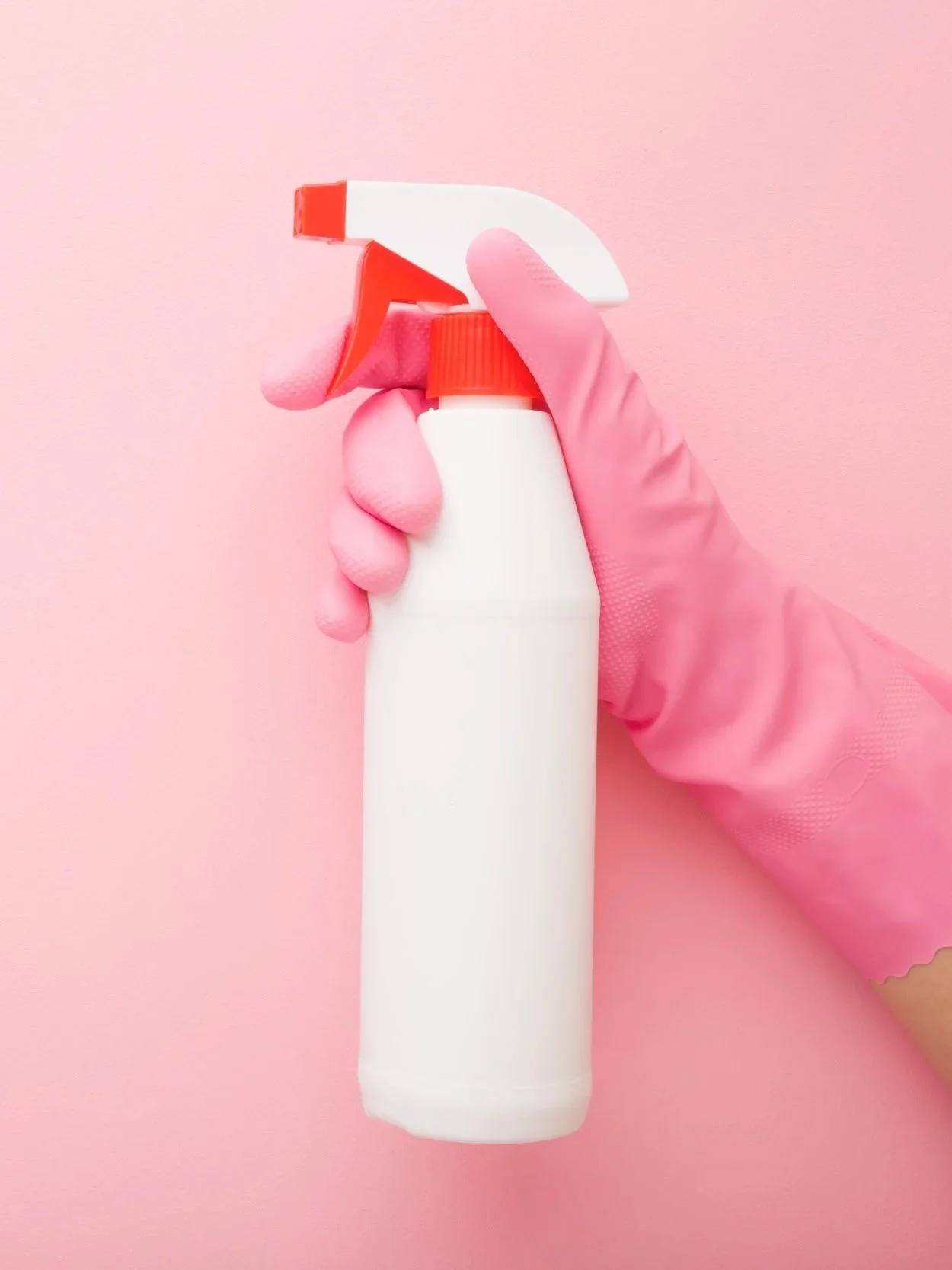 A person wearing pink rubber gloves holding a white spray bottle with a red and white nozzle against a pink background.