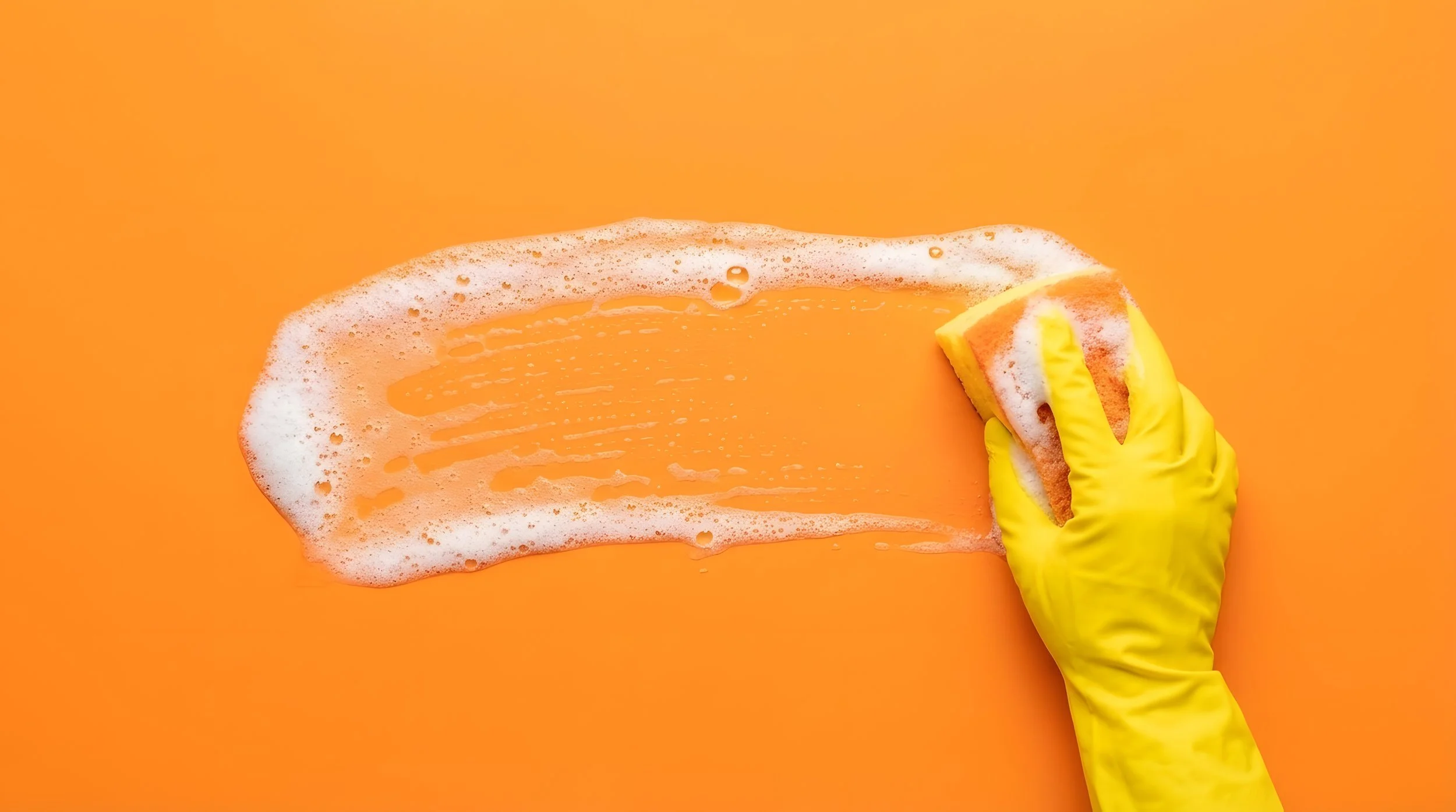 A person wearing a yellow rubber glove cleaning a bright orange surface with a sponge, producing soap suds and cleaning streaks.