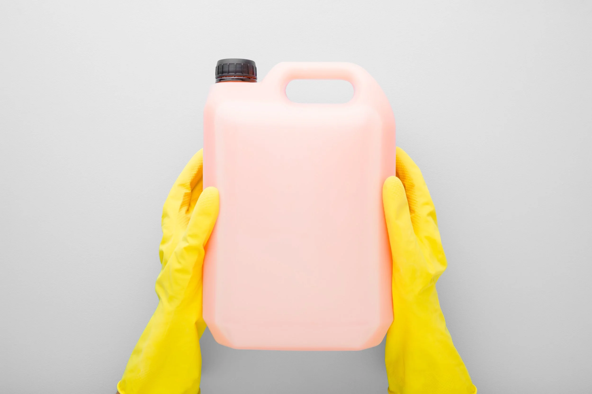 Person wearing yellow gloves holding a pink plastic jug with a black cap against a gray background.