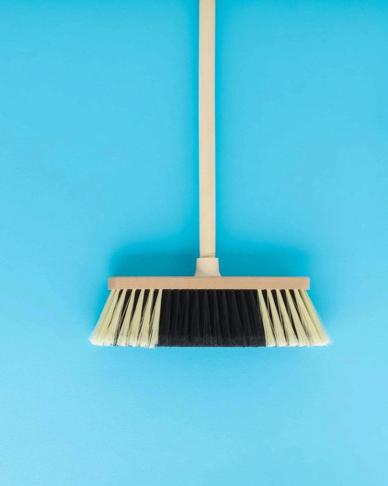 A wall-mounted broom with a wooden handle and bristles in black and cream, hanging against a bright blue wall.