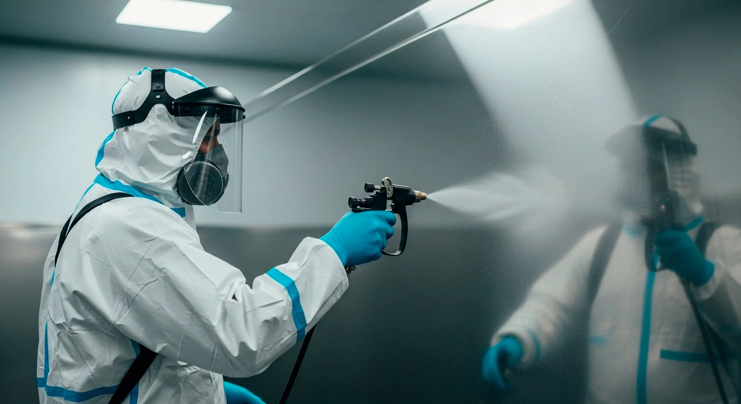 A person in full protective gear, including a hazmat suit, face shield, and gloves, is using a spray gun to disinfect a metallic surface in a controlled environment.
