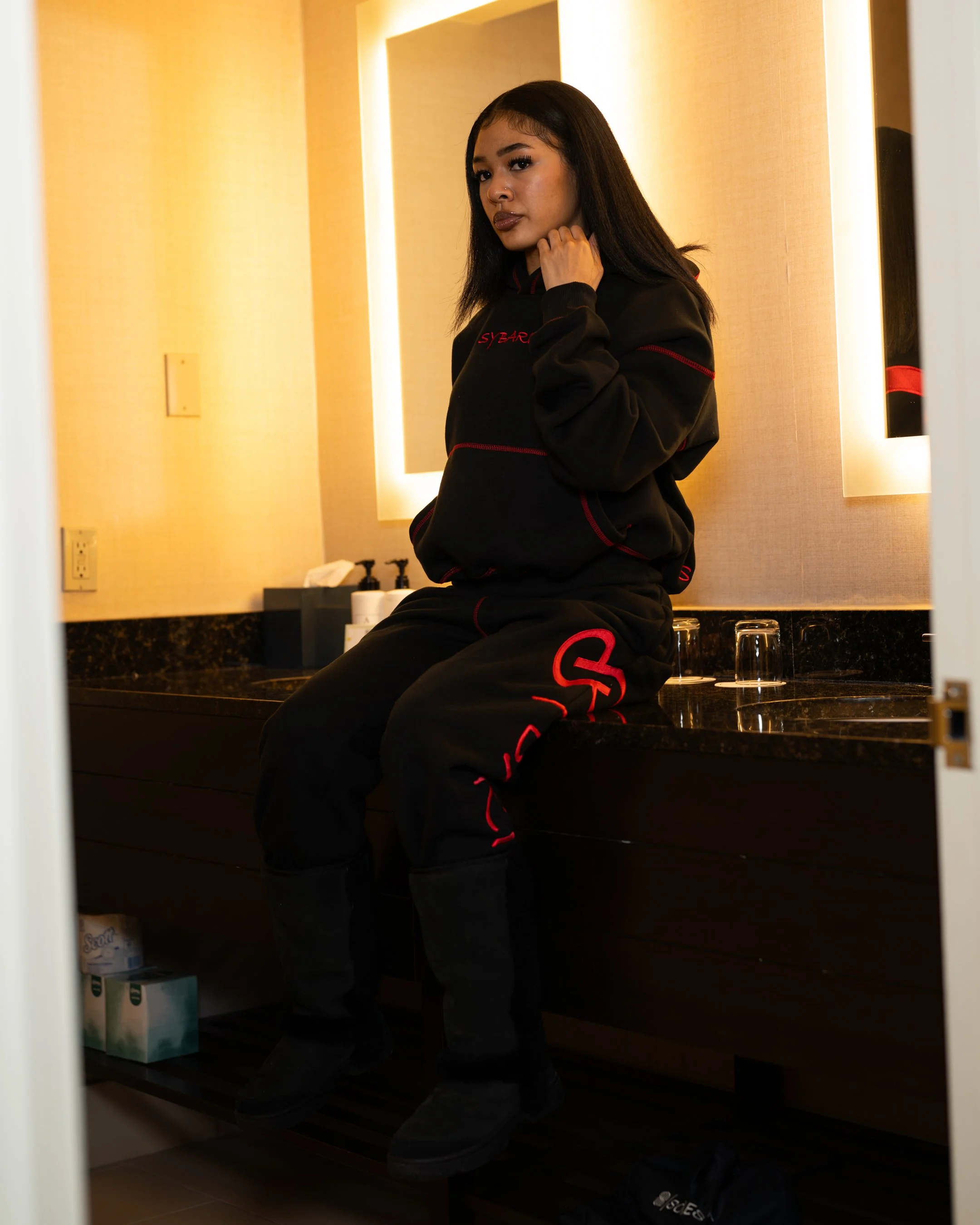 Young woman sitting on bathroom counter in black and red casual outfit, looking at the camera with a serious expression, illuminated mirrors behind her.