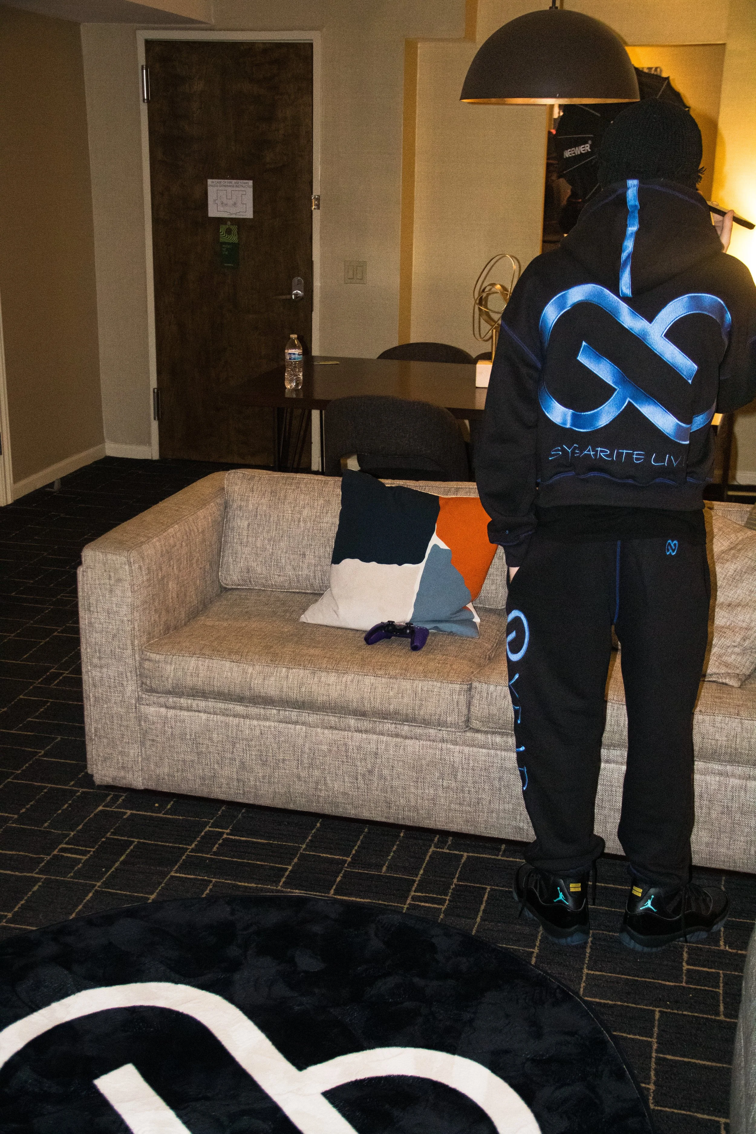 A person standing in a hotel room wearing black streetwear with blue accents, holding a microphone, with a beige sofa, colorful pillow, and a gaming controller on the sofa. There is a dining table with a water bottle and a lamp hanging above.